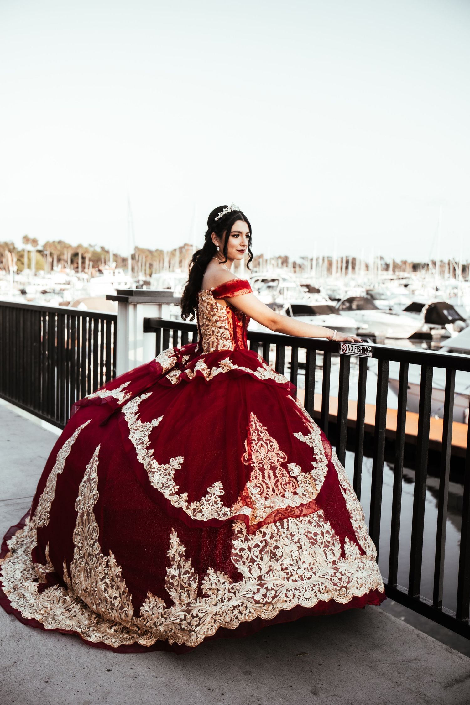 Young woman in stunning red and gold ball gown poses on marina waterfront balcony.