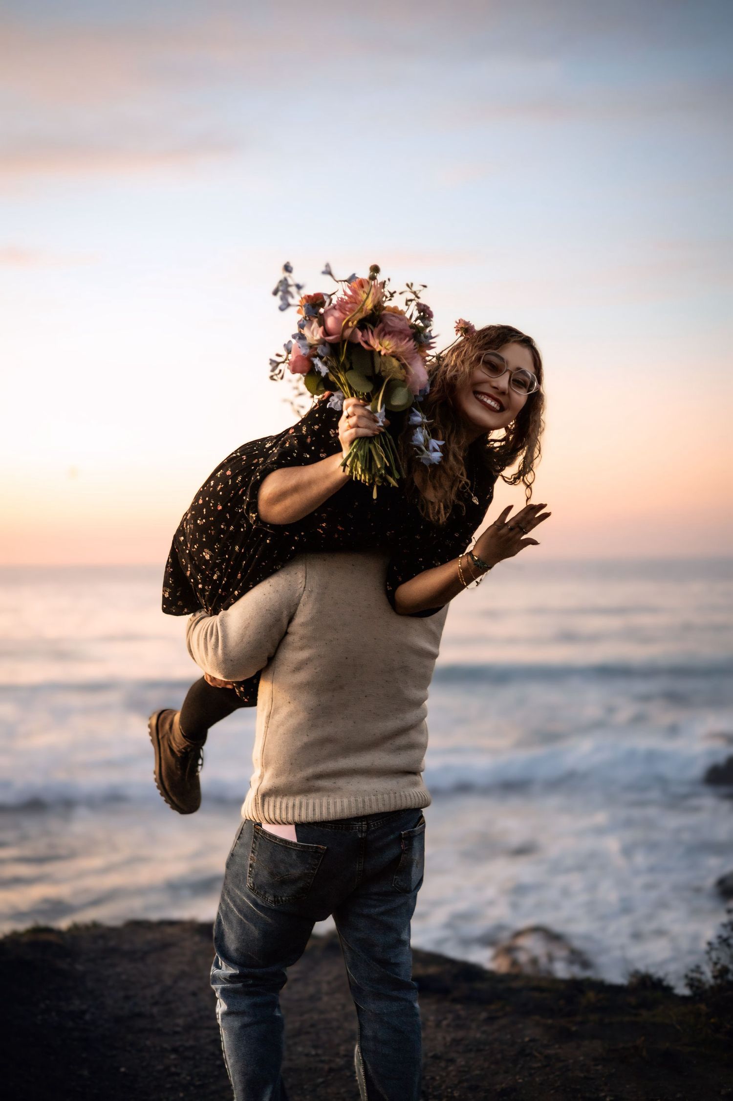 A man carries a laughing woman holding a flower bouquet on his back at a stunning ocean sunset.