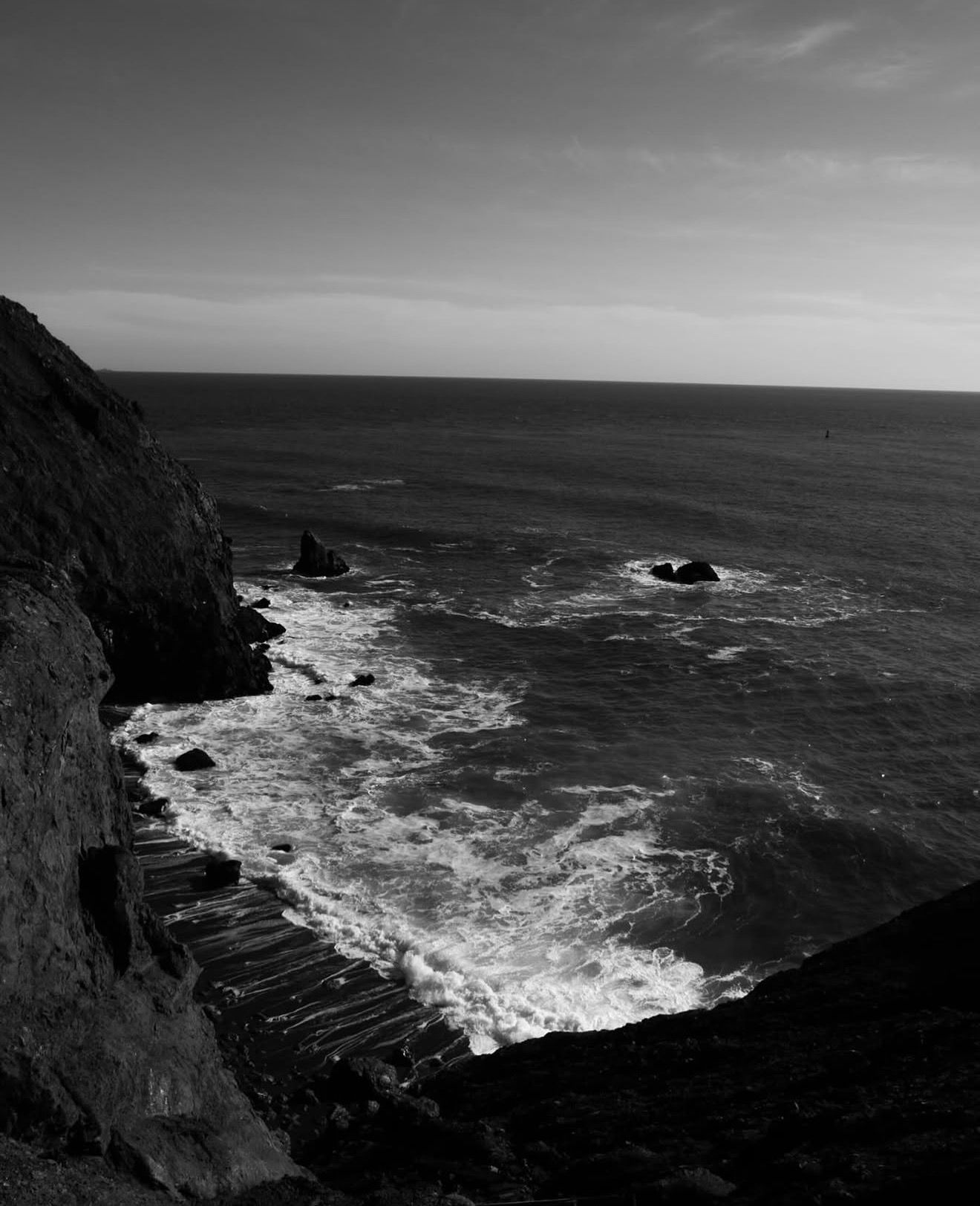 Black and white coastal seascape with rocky cliffs, crashing waves, and sea stacks along a dramatic ocean shoreline.