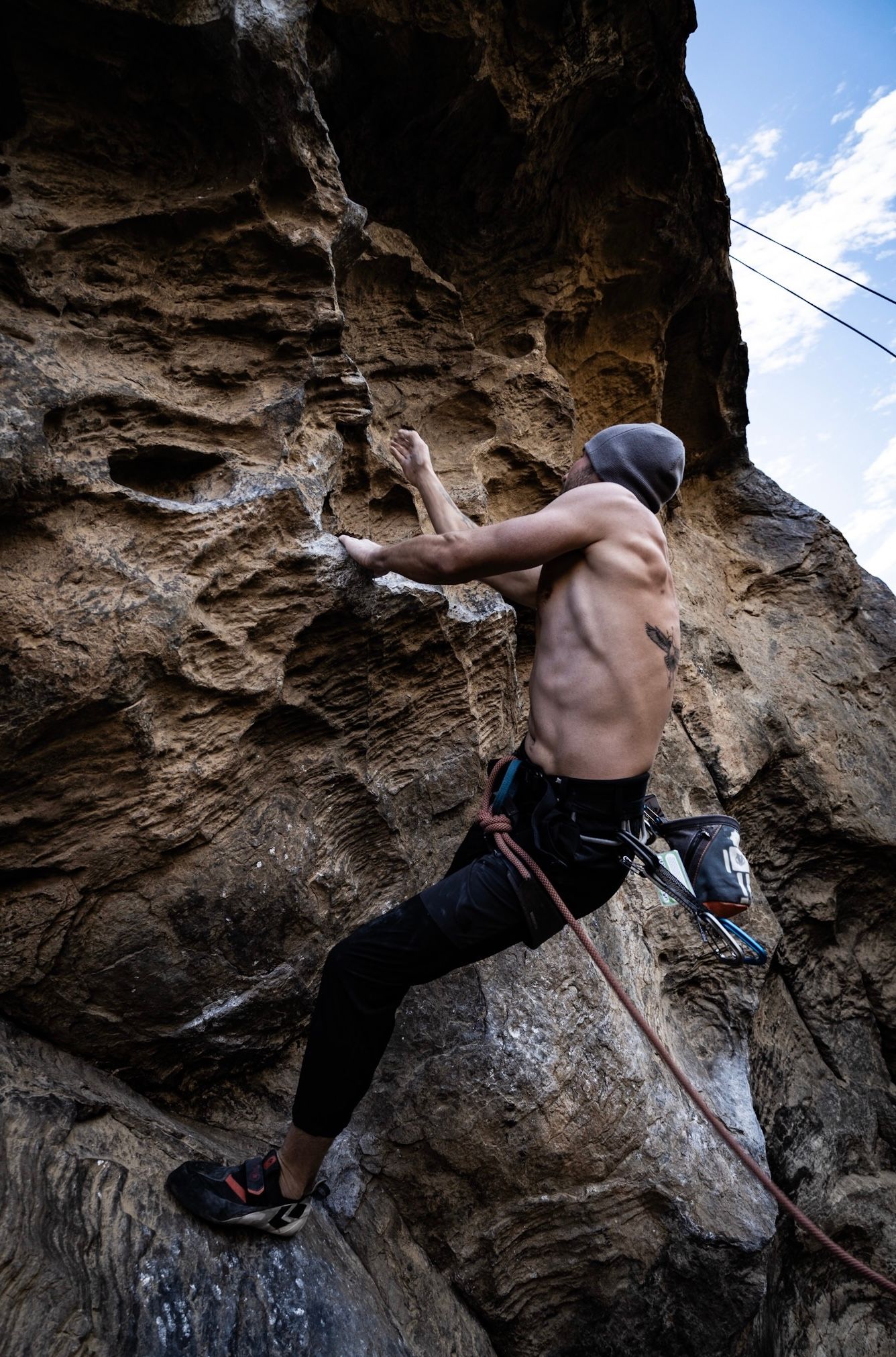 Shirtless male rock climber scaling a rugged brown cliff face with rope safety gear against a cloudy sky.
