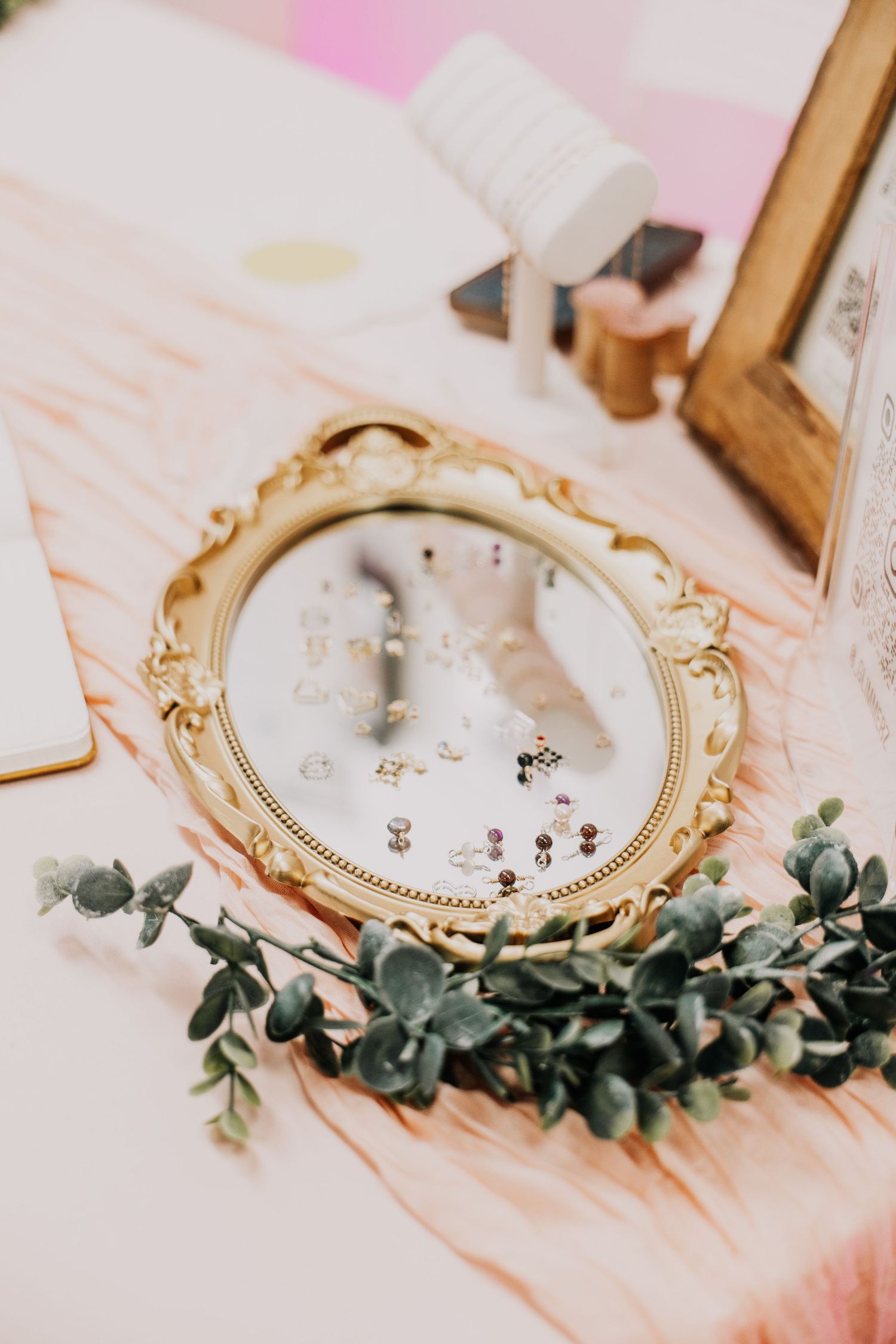 Ornate gold oval mirror displaying earring collection on pink marble table with eucalyptus greenery.