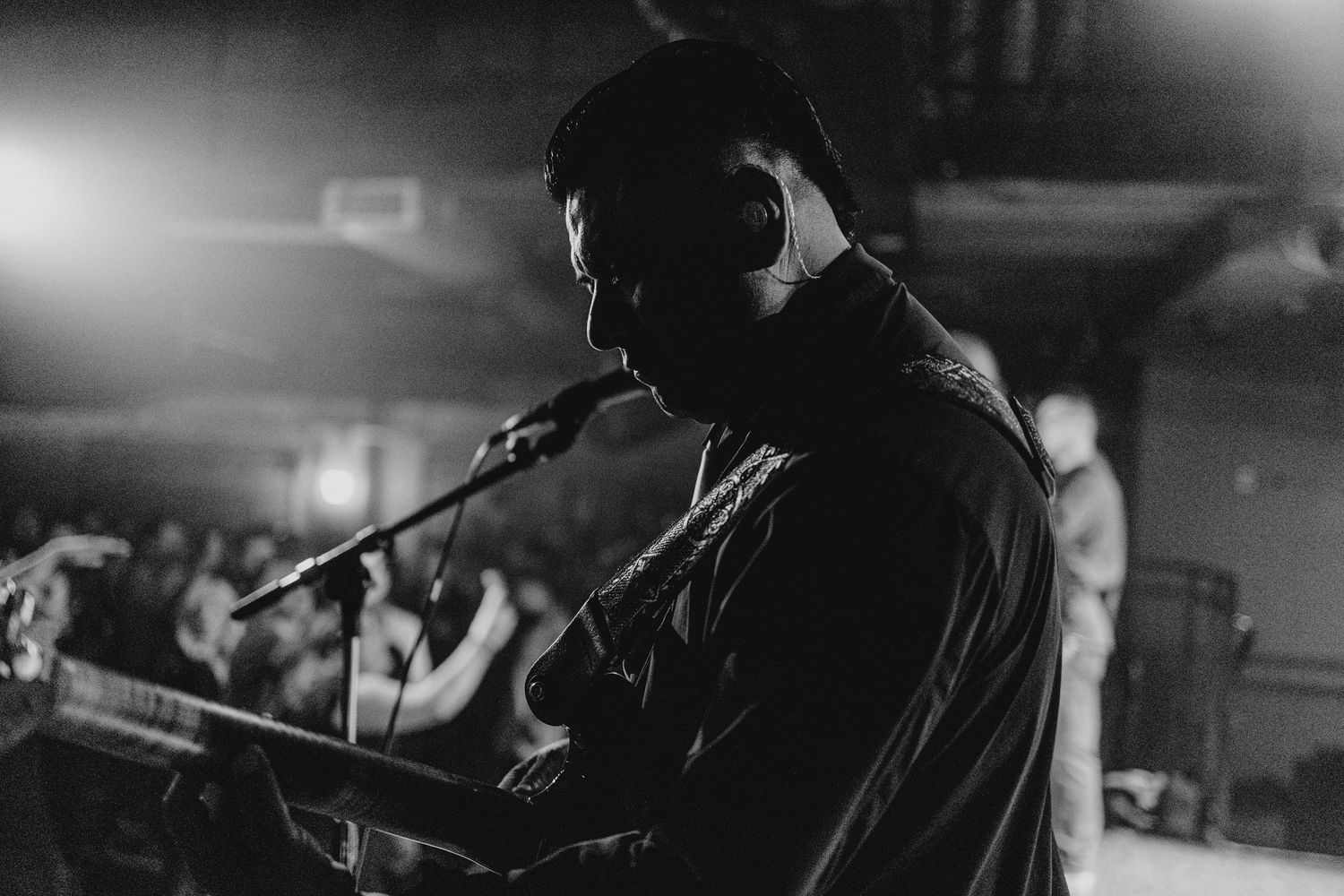 Black and white side profile of a musician singing into a microphone with headphones at a live concert.
