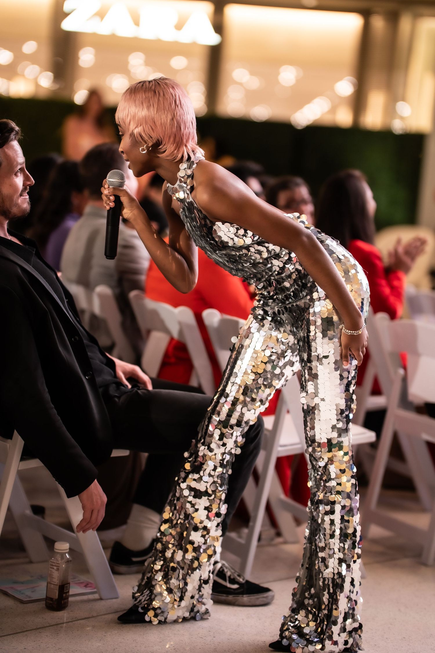 Performer in a sequined jumpsuit singing with a microphone to a seated audience at an evening event.