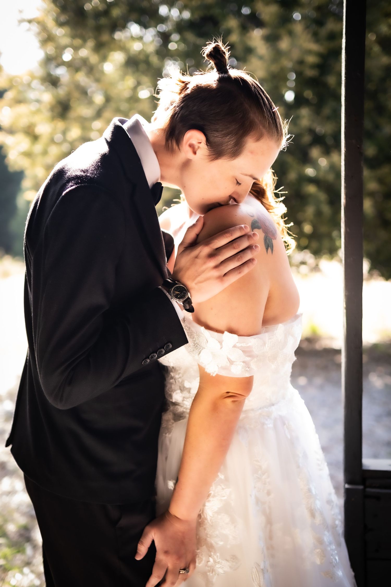 Groom in black suit kissing bride on shoulder, bride in off-shoulder white gown outdoors.