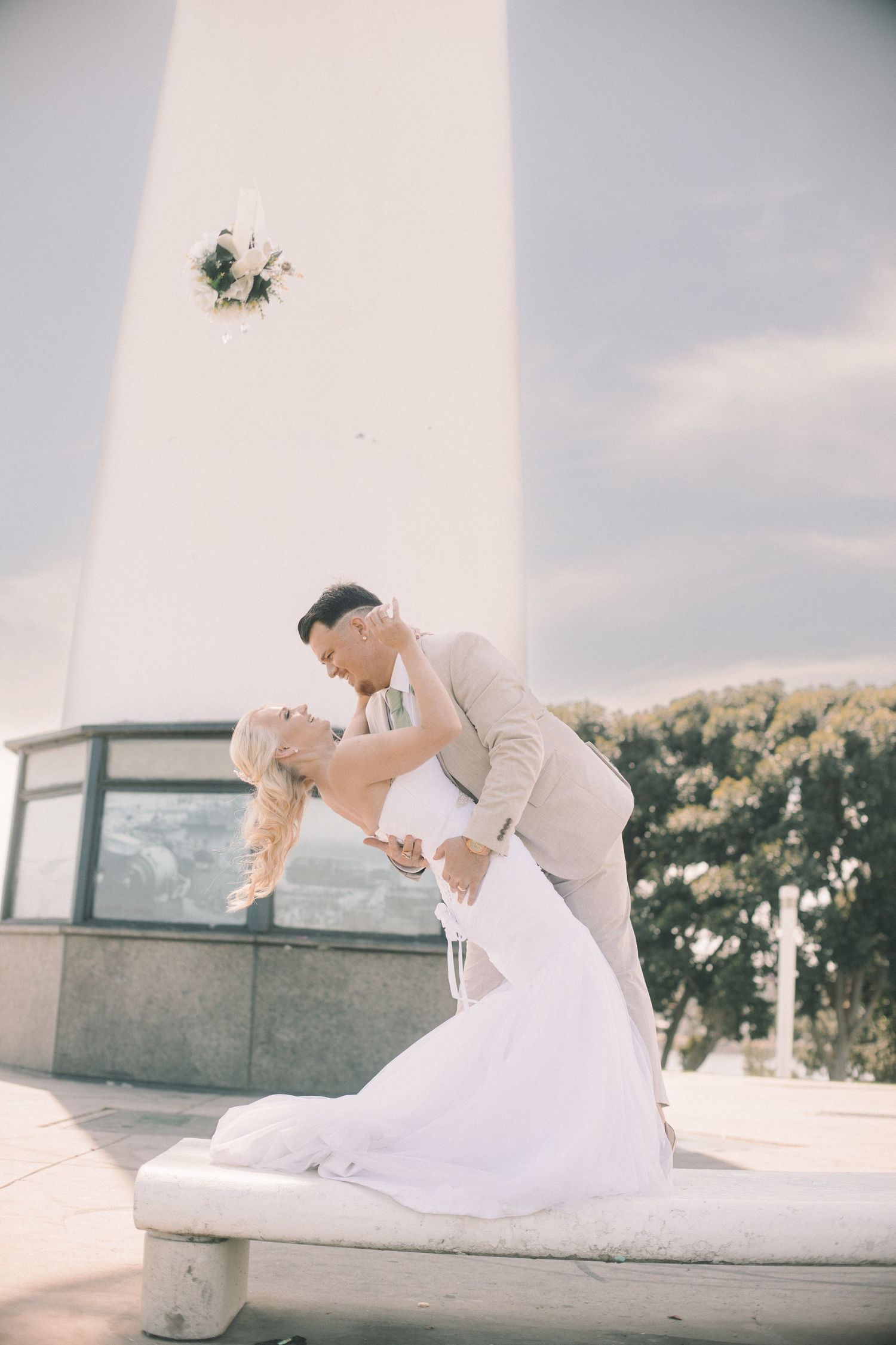 A groom dips his bride for a kiss in front of a tall white lighthouse outdoors.