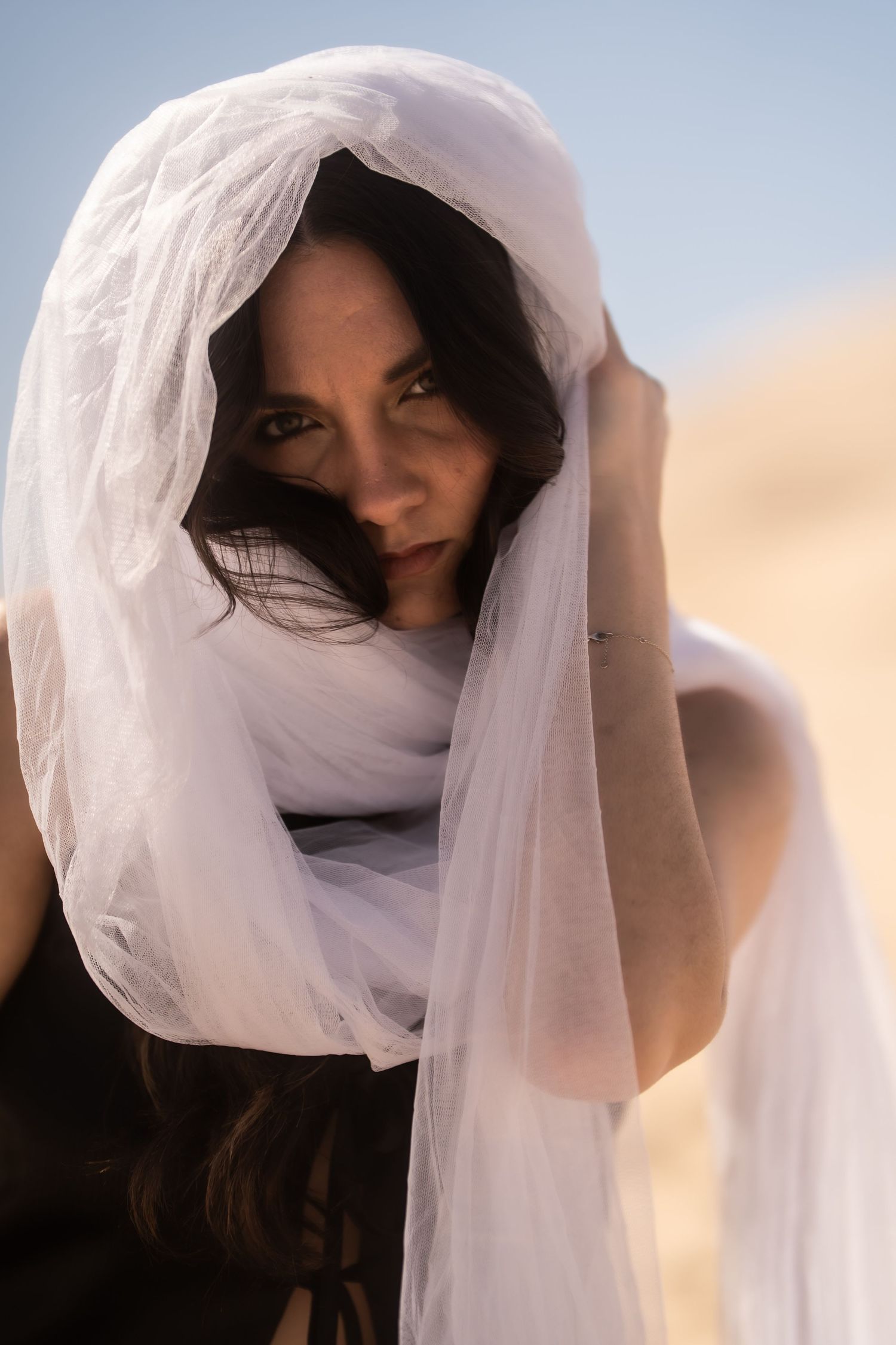Dark-haired woman wearing a flowing white veil headscarf in a desert setting gazing at the camera.