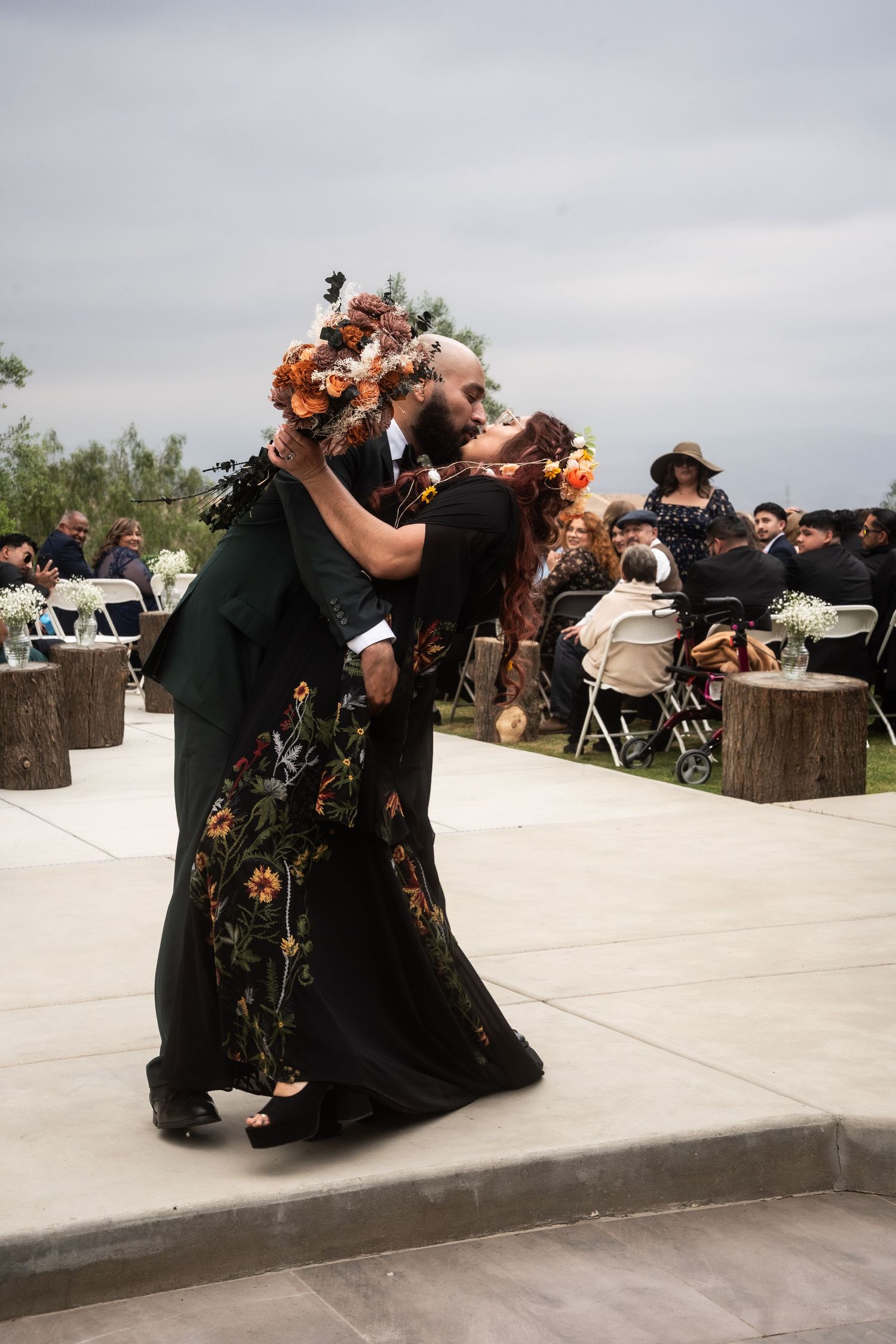 Couple sharing a first kiss in black floral attire with flower crowns at an outdoor wedding ceremony.