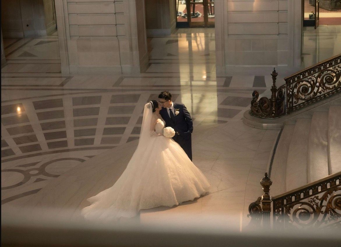 Bride and groom share romantic kiss in grand marble hall with elegant staircase.