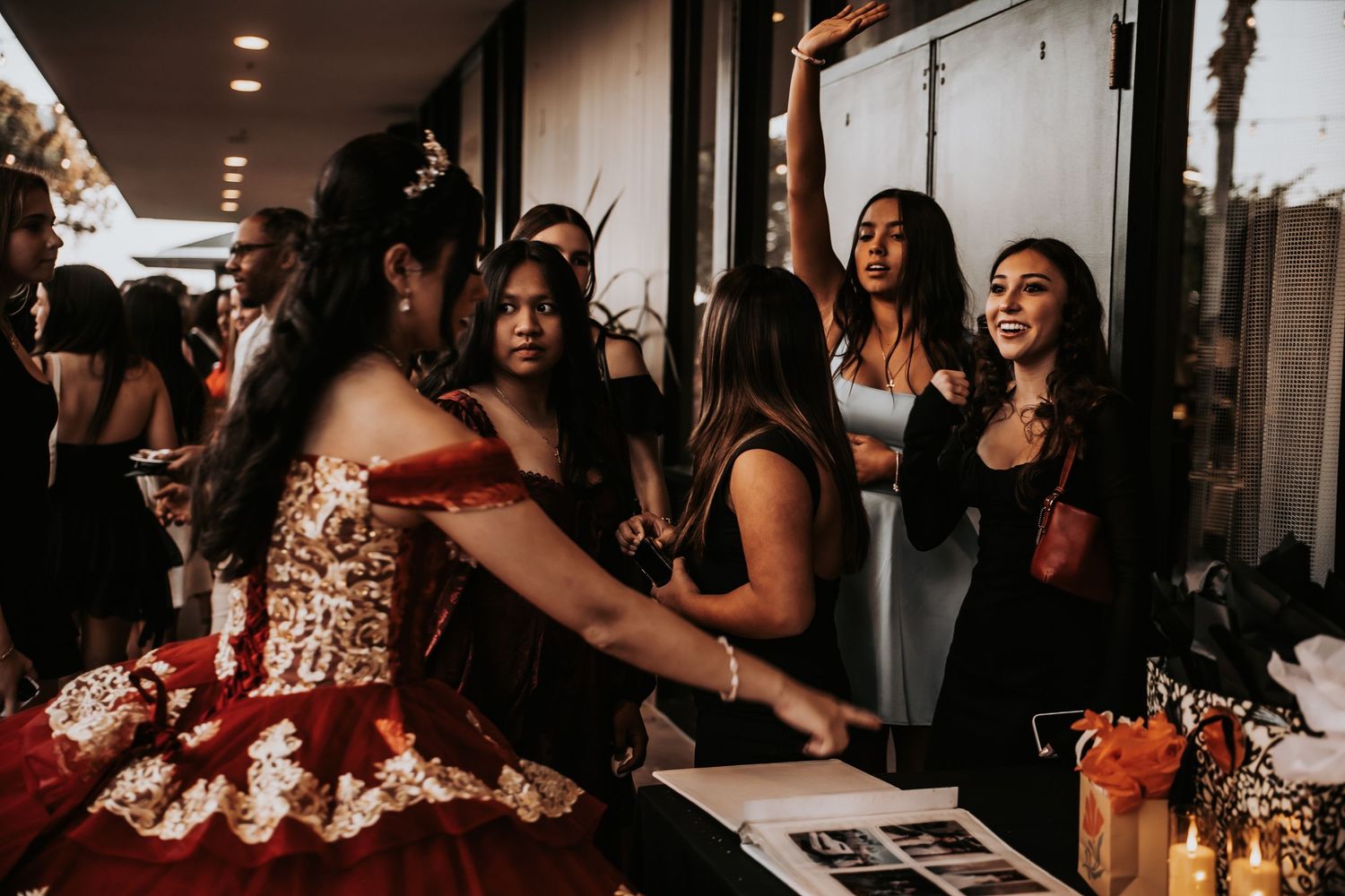 Quinceañera girl in red gold gown laughs with friends at festive party celebration indoors.
