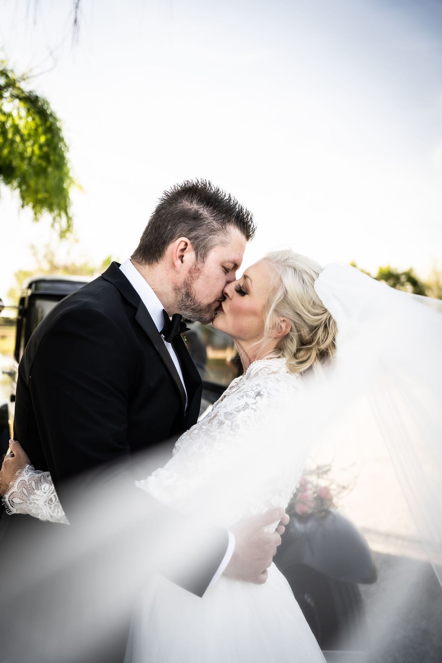 A bride and groom share a passionate kiss outdoors, with the veil flowing in the wind.