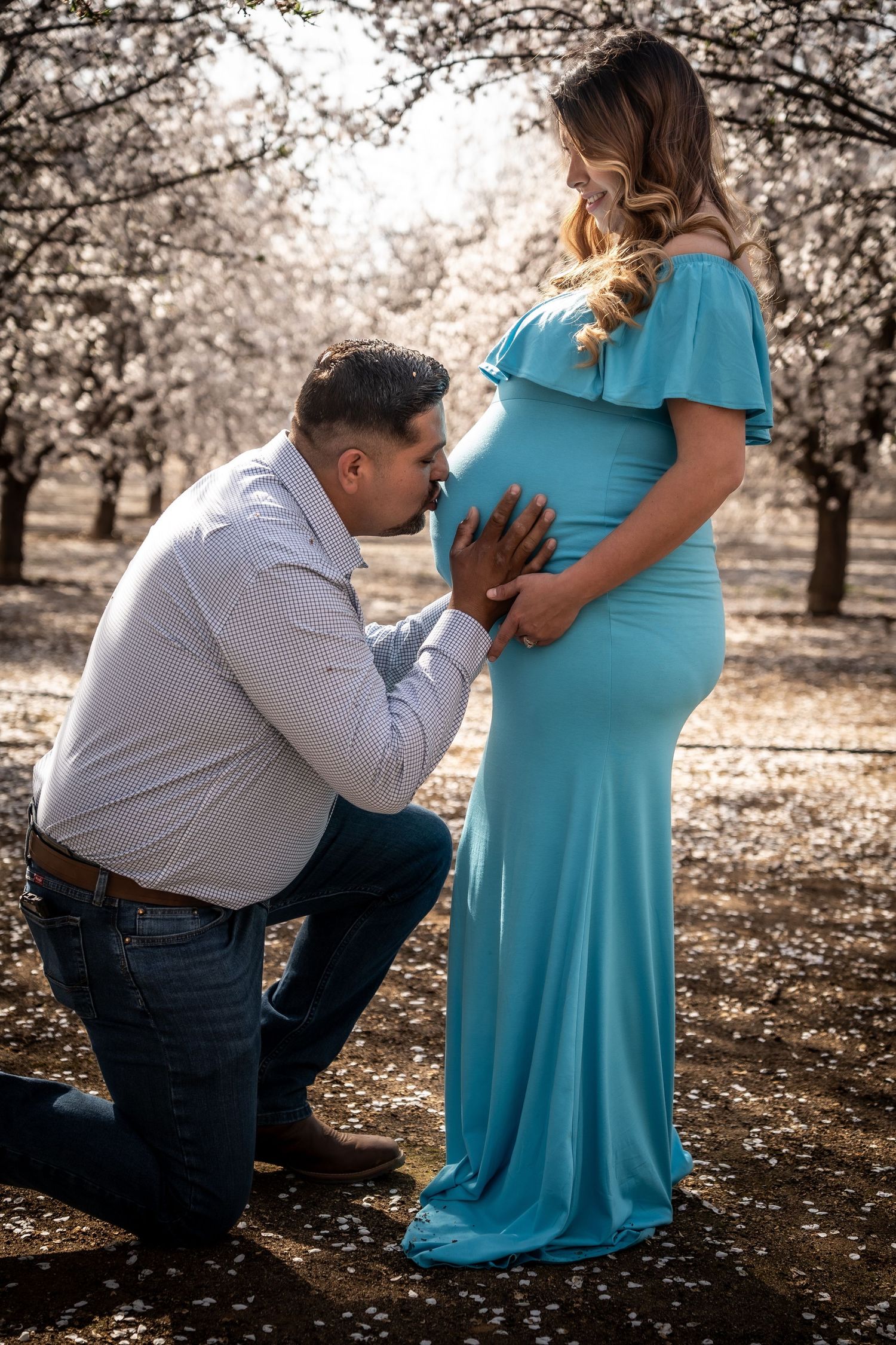 A man kneels to kiss a pregnant womans belly in a teal gown among blooming trees.