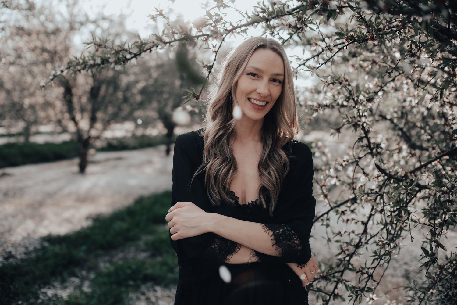 Happy blonde woman in black blazer posing with arms crossed near spring blossoming trees.