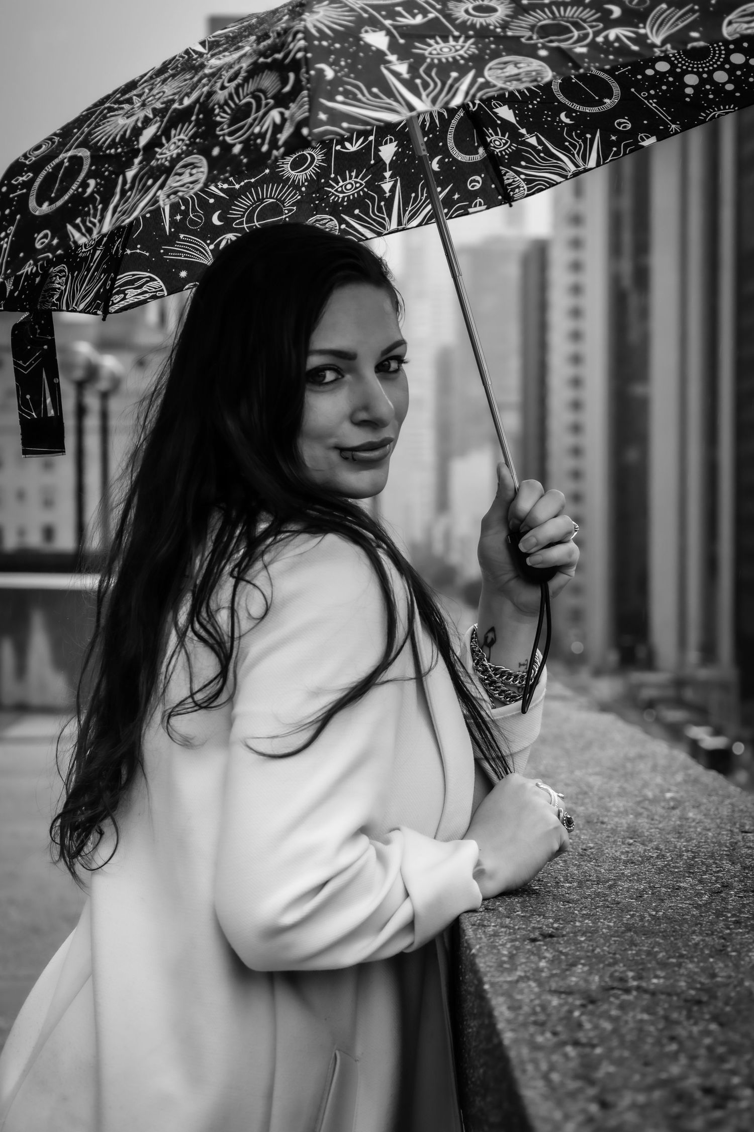 A woman in a white coat holding a patterned umbrella in a black and white urban portrait.