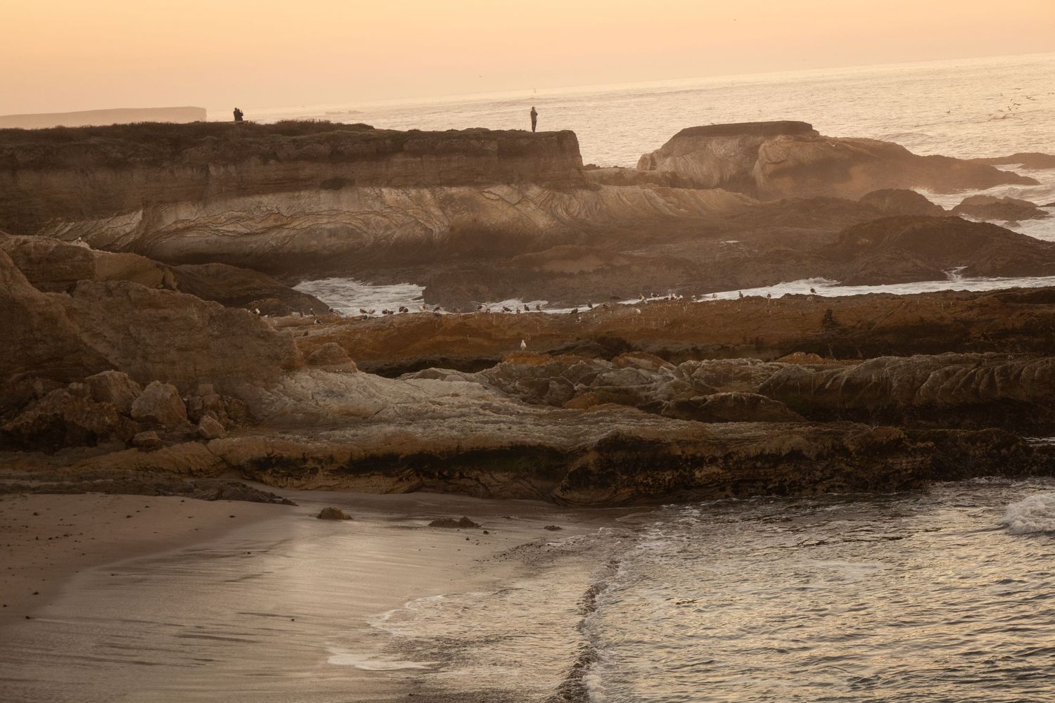 Rocky coastal cliffs at sunset with waves gently lapping the shore and silhouettes of people on the rocks.