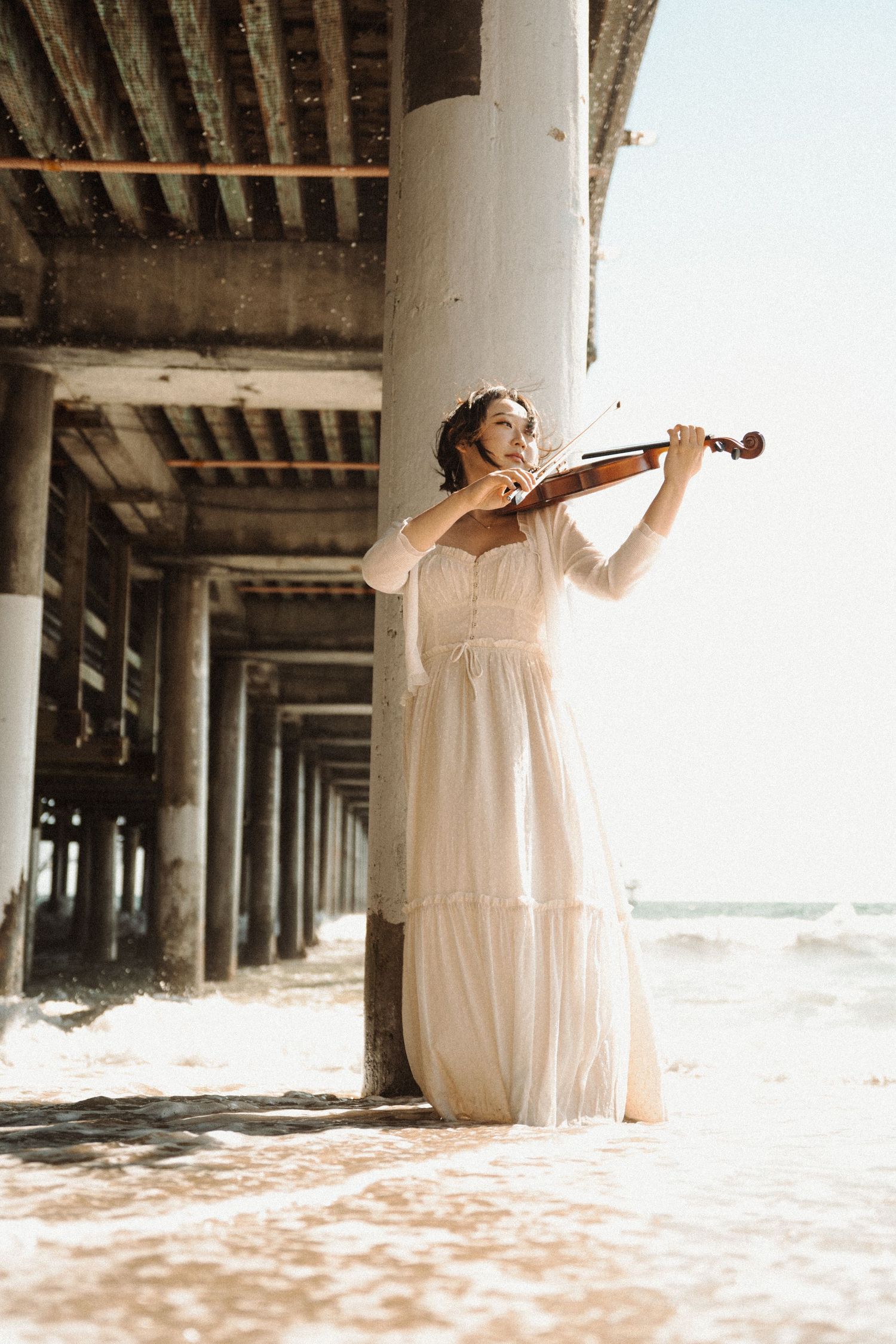 Woman in flowing white dress playing violin beside a beach pier column.