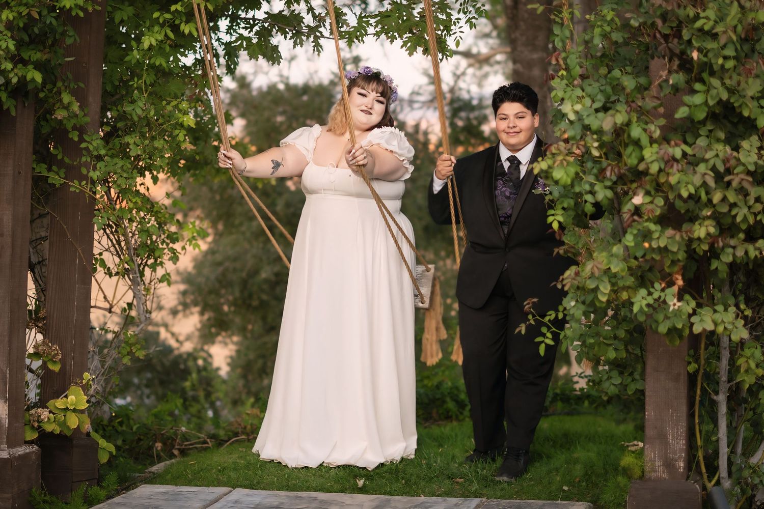Bride in white gown and groom in black suit posing on a wooden swing surrounded by lush green ivy outdoors.