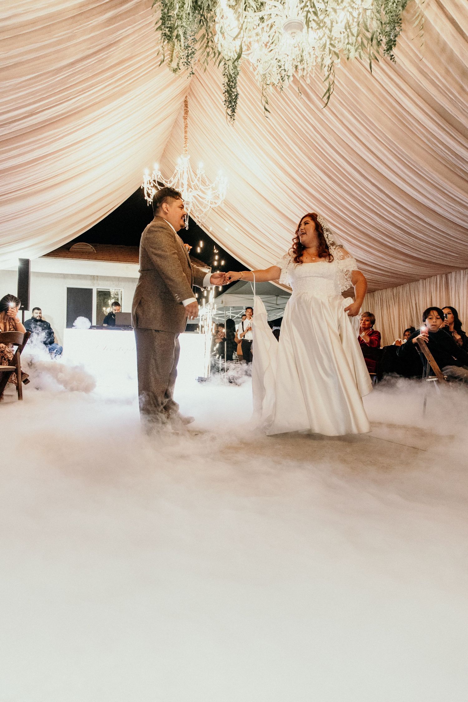 Bride and groom share first dance on fog-covered floor beneath chandelier in draped reception tent.