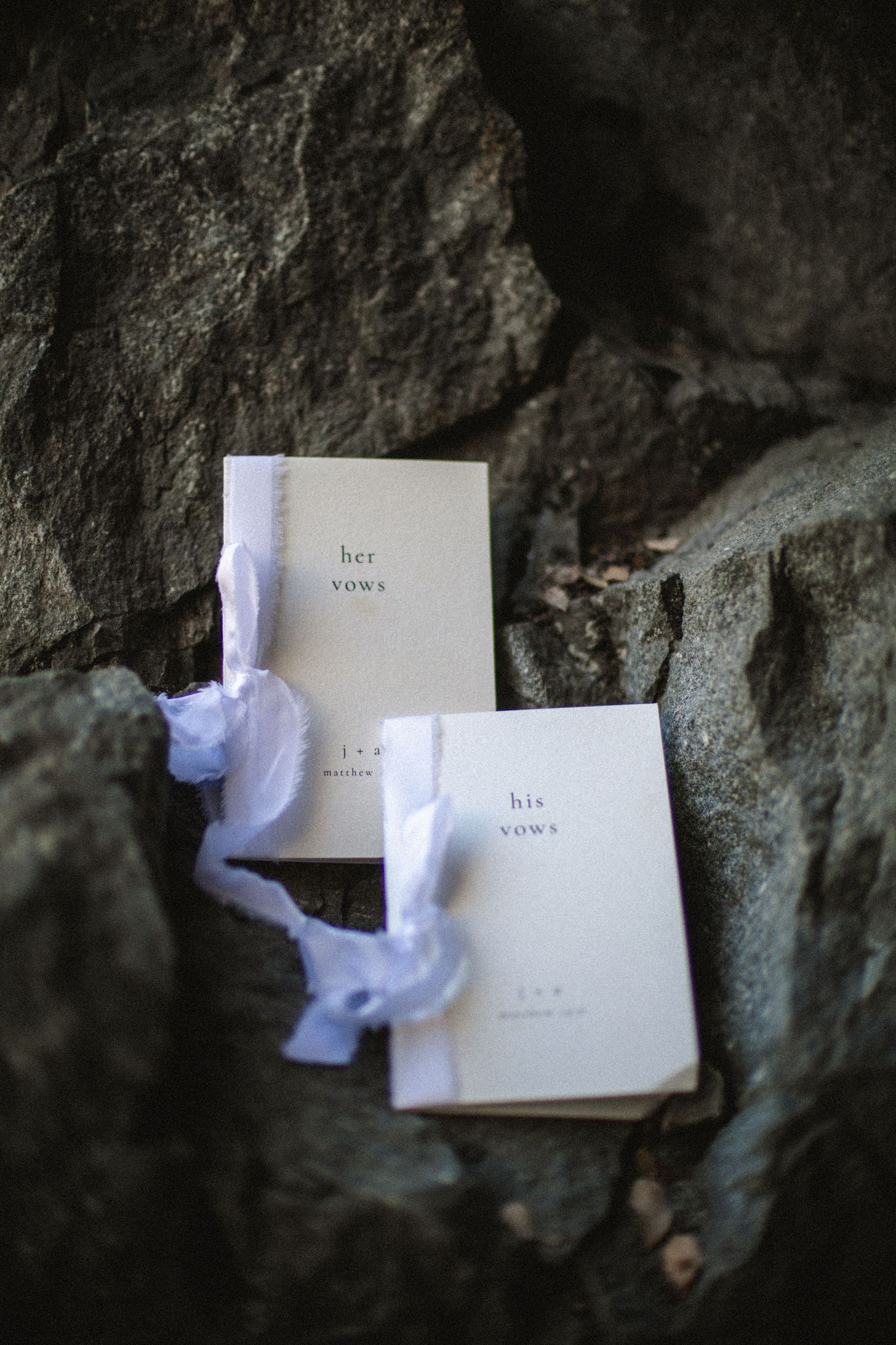 Two wedding vow booklets labeled his vows and her vows resting on rustic rocks with white ribbon.