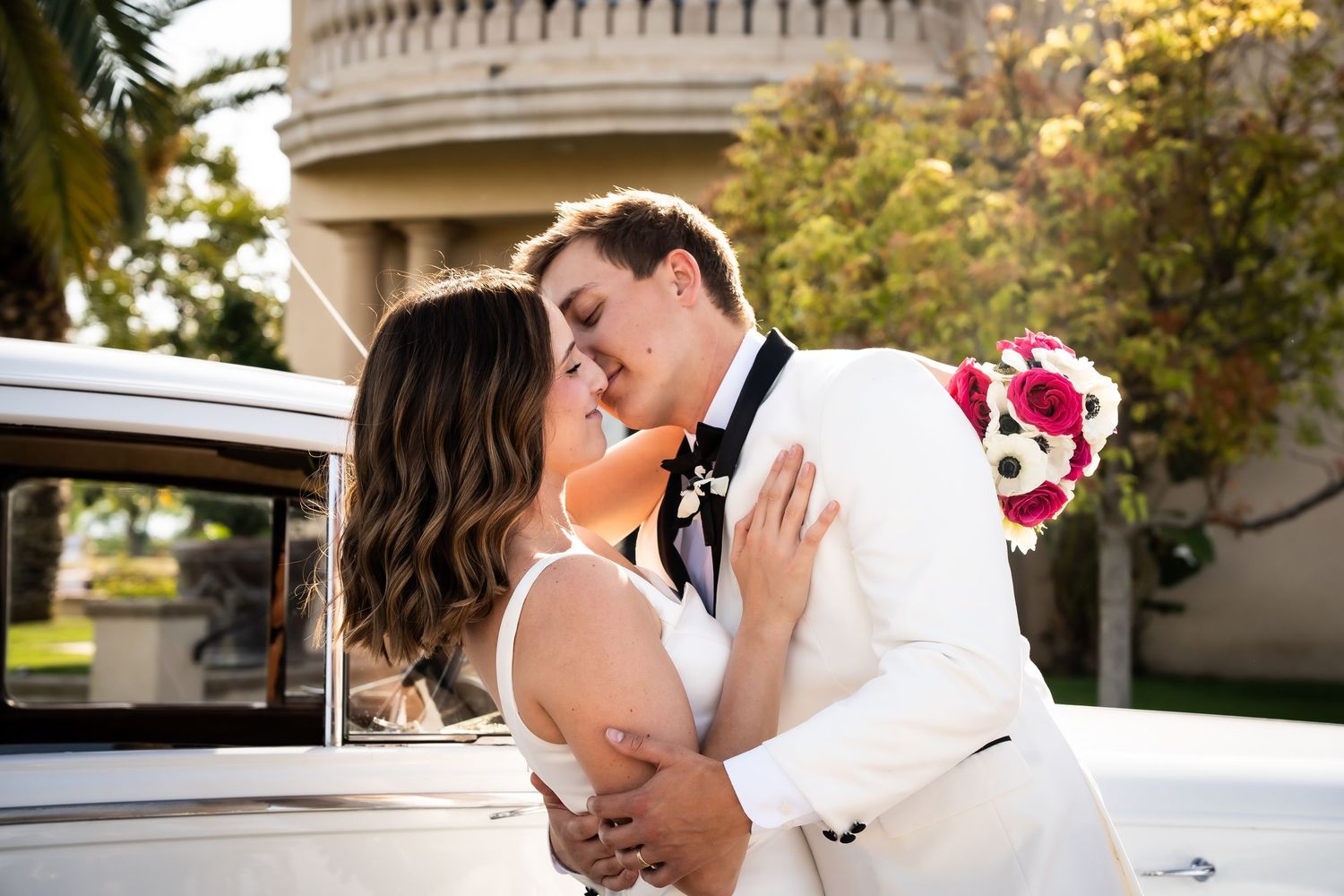 Newlyweds sharing a kiss beside a vintage white car with a bridal bouquet.