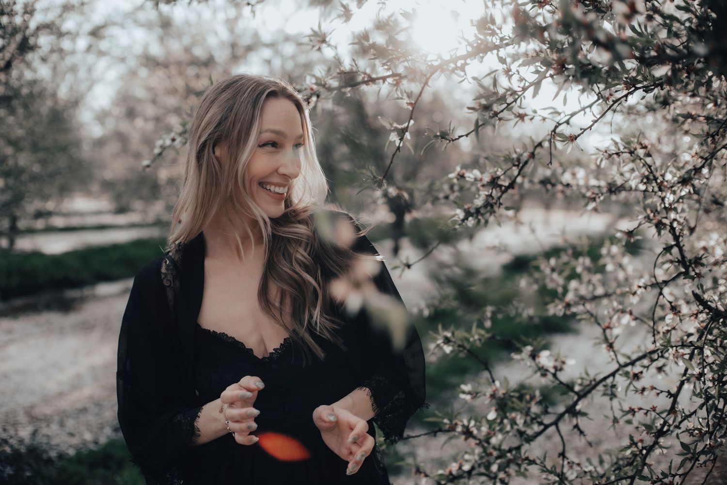 Smiling woman in black outfit standing near blooming white cherry blossom trees outdoors.