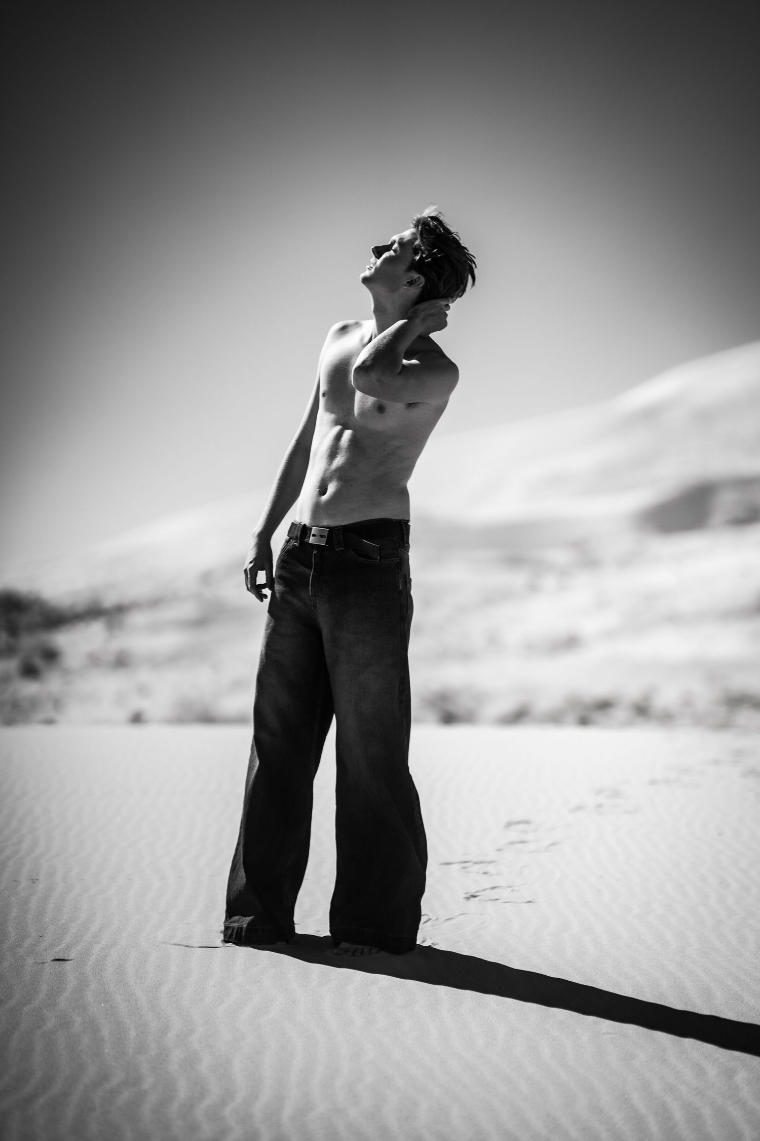 Shirtless athletic man standing on sand dunes looking up at the sky in a black and white desert photo.