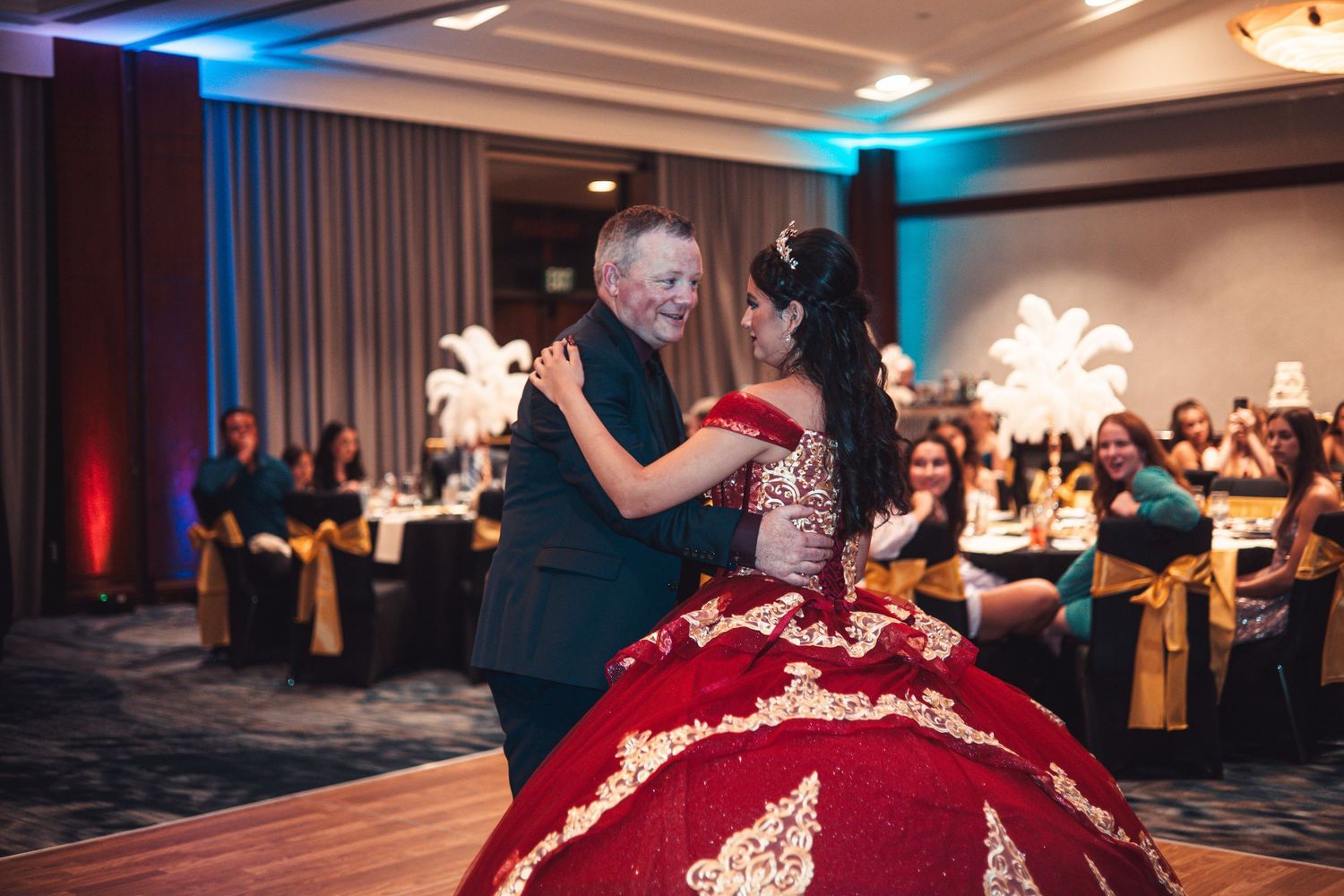 Young woman in red and gold quinceañera gown dances with older man at elegant ballroom celebration.