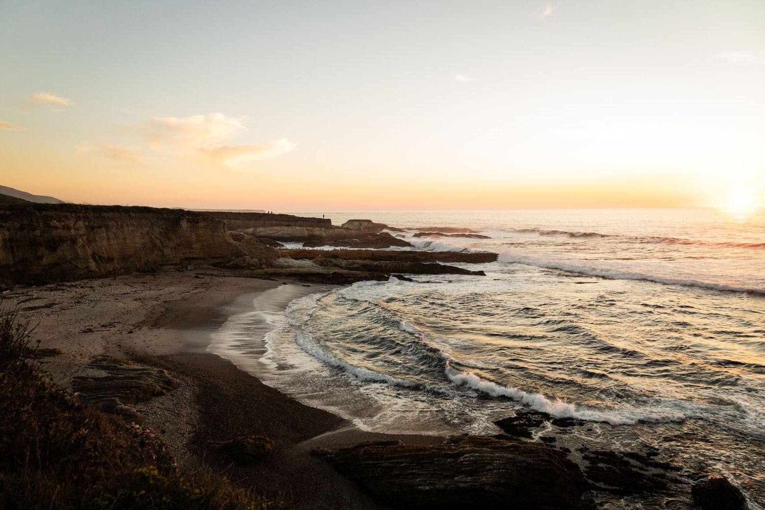 Golden sunset over a rocky coastal beach with gentle ocean waves and dramatic cliffs along the shoreline.