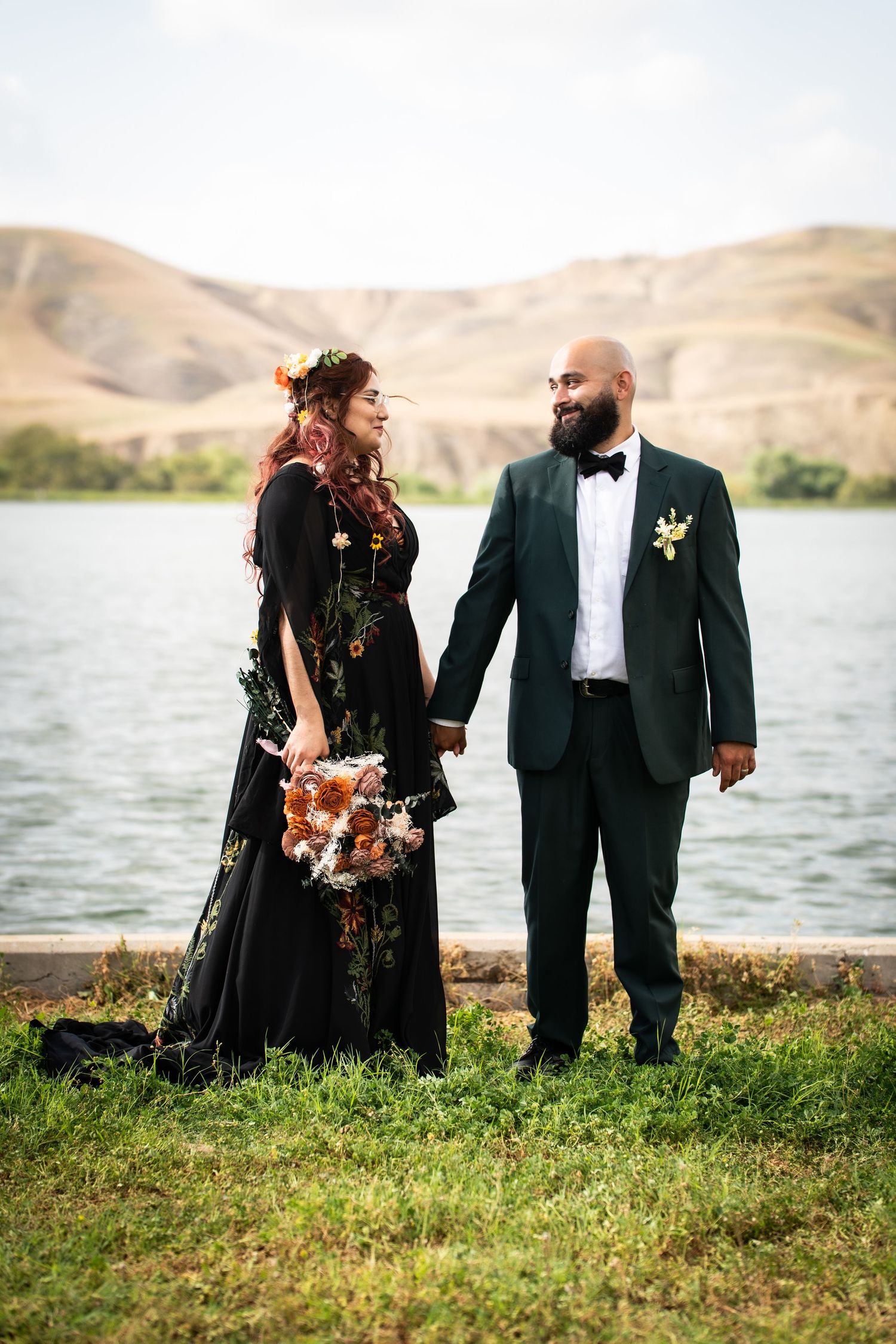 Bride in black gown and groom in dark green tuxedo hold hands by a lake with hills in the background.