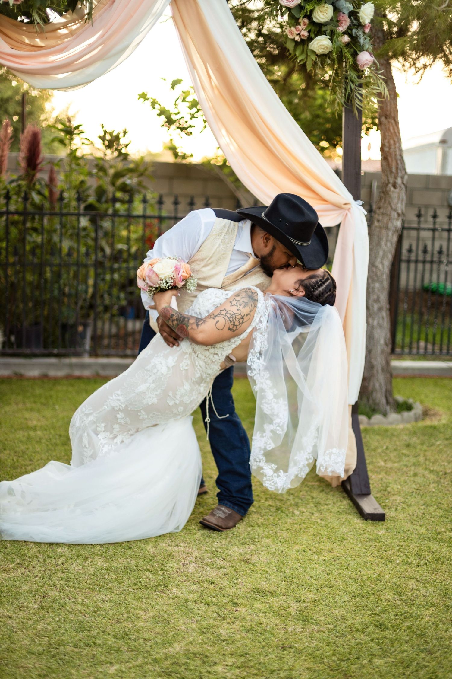 Bride and groom share a romantic dip kiss under a floral arch, groom wearing a black cowboy hat at outdoor wedding.