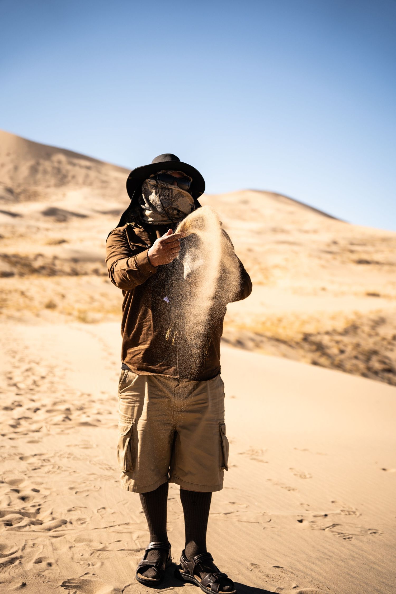 Man in hat and face mask pouring sand in a desert landscape.