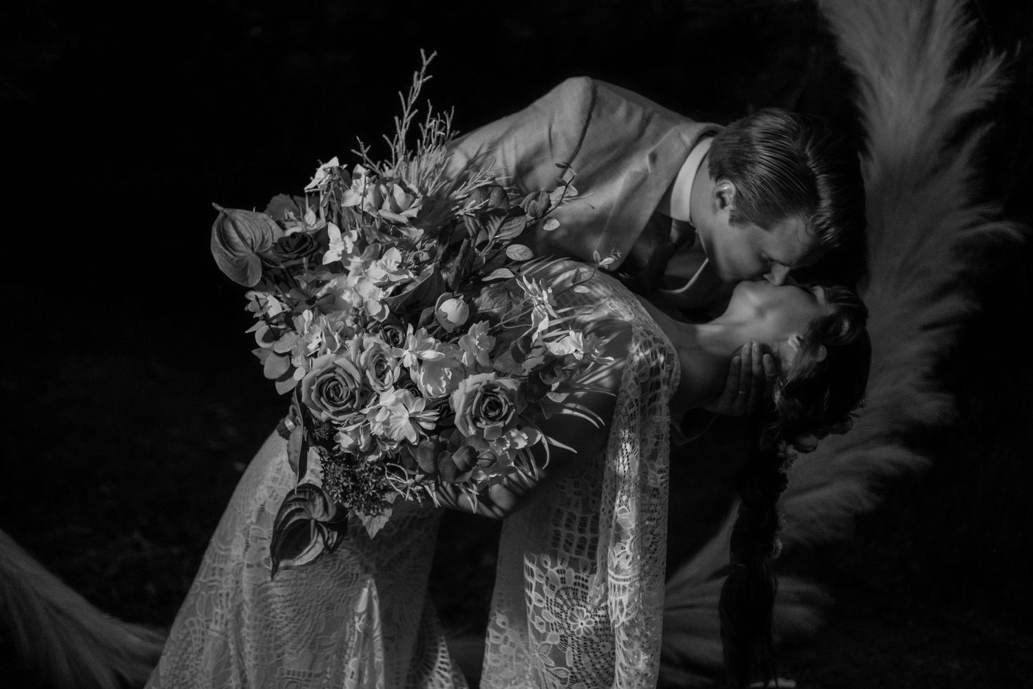 Bride and groom sharing a romantic dip kiss with a lush floral bouquet in black and white.