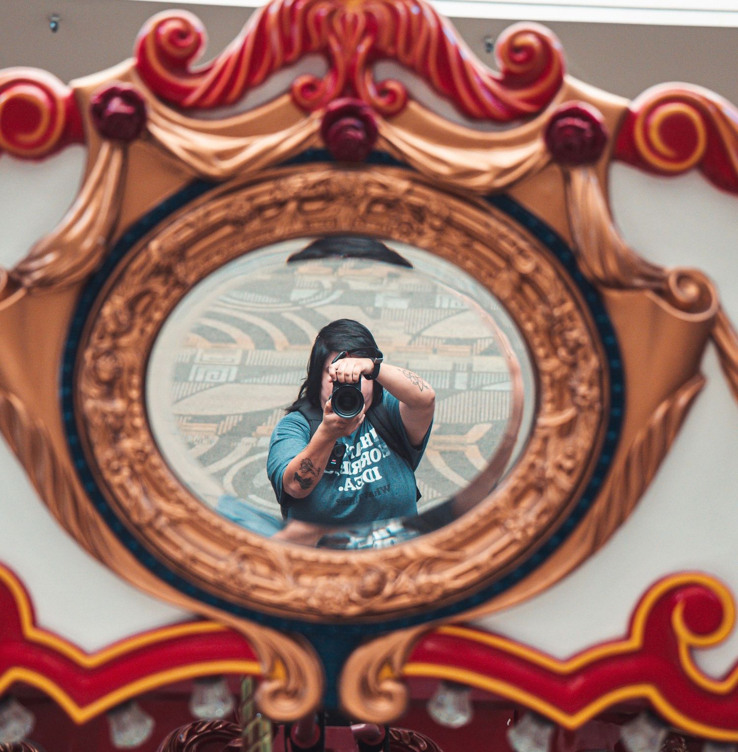 Photographer taking selfie reflected in decorative oval mirror of vibrant red and gold carousel.