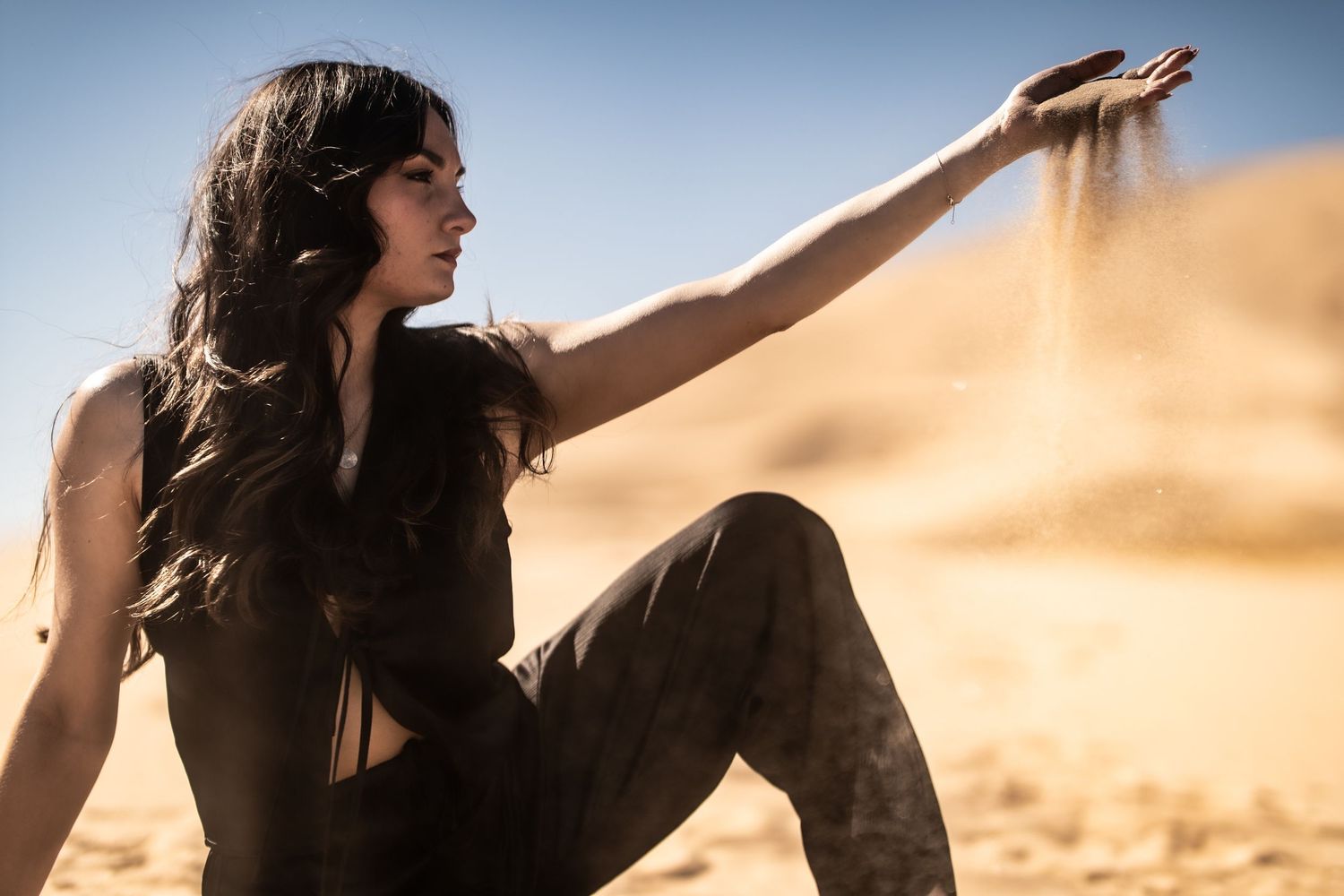 Woman sitting on desert sand dunes letting sand fall from her outstretched hand in bright sunlight.