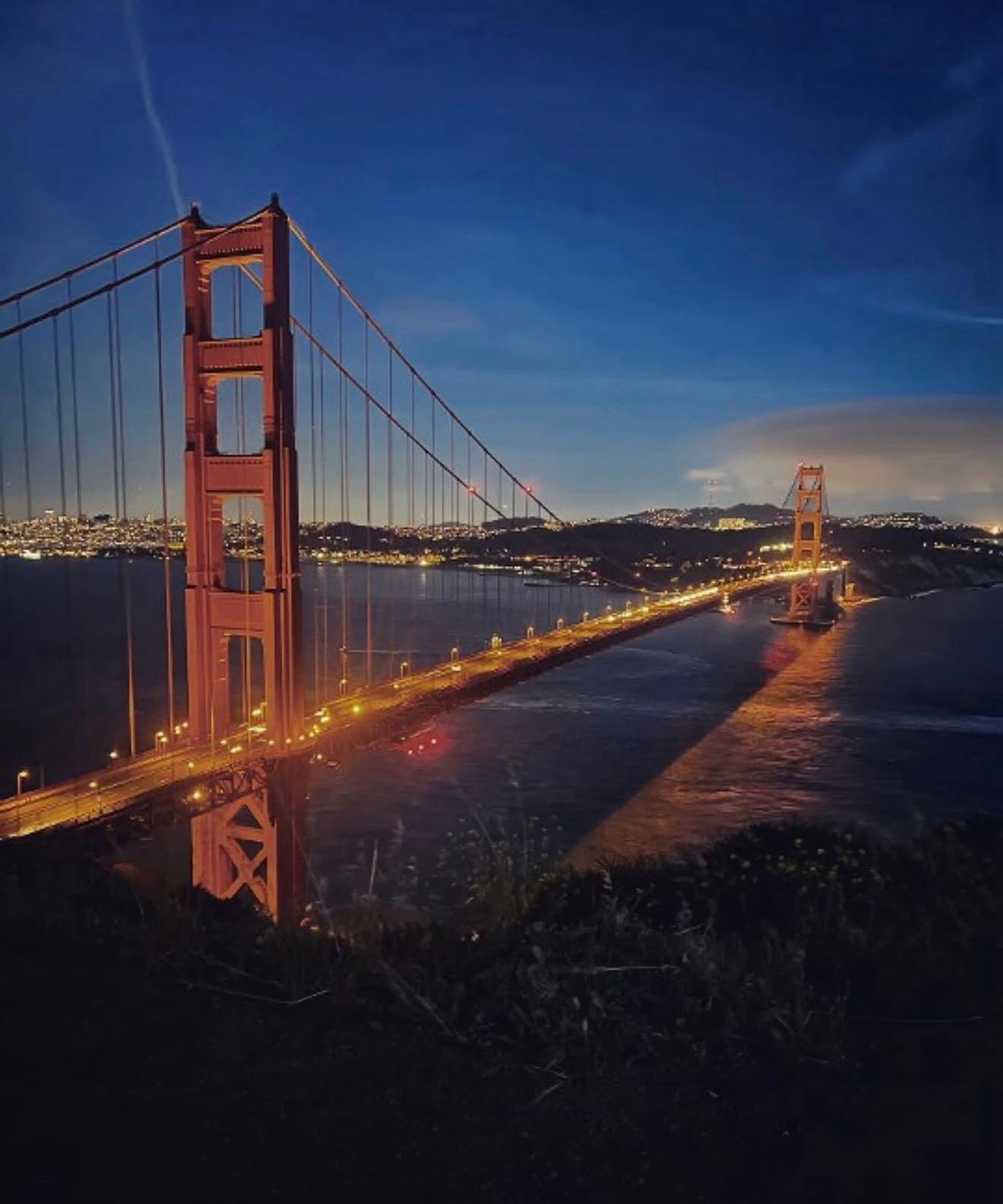 Golden Gate Bridge illuminated at night with city lights reflecting on San Francisco Bay under a deep blue twilight sky.