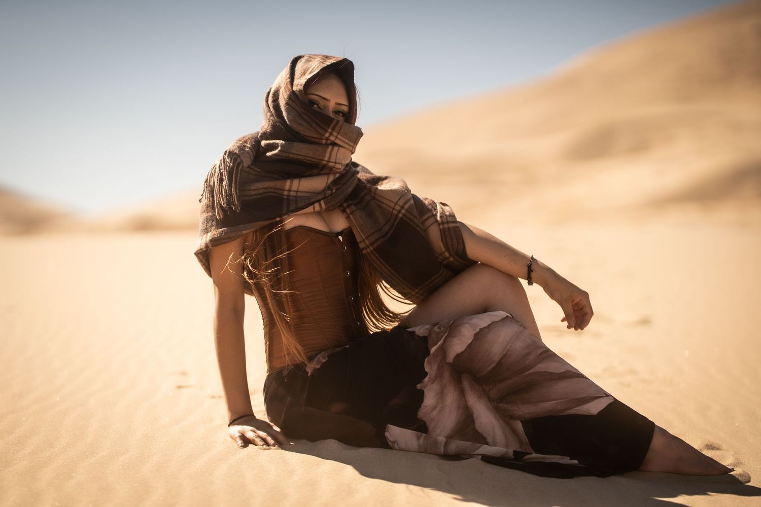 Woman in desert nomad outfit sitting on sand dunes in bright sunlight.