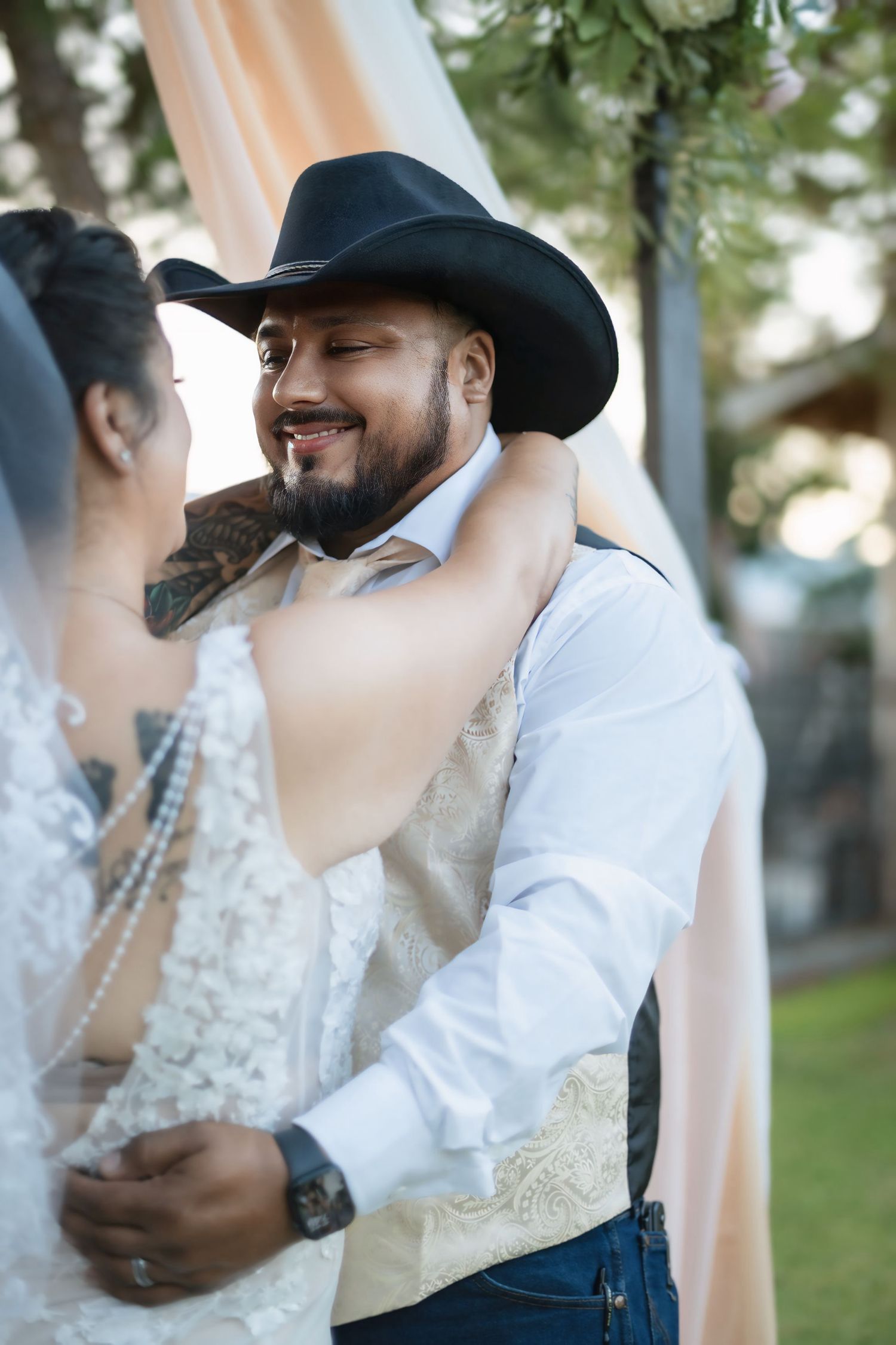 Groom in black cowboy hat and vest smiles while sharing first dance with bride in lace wedding dress outdoors.