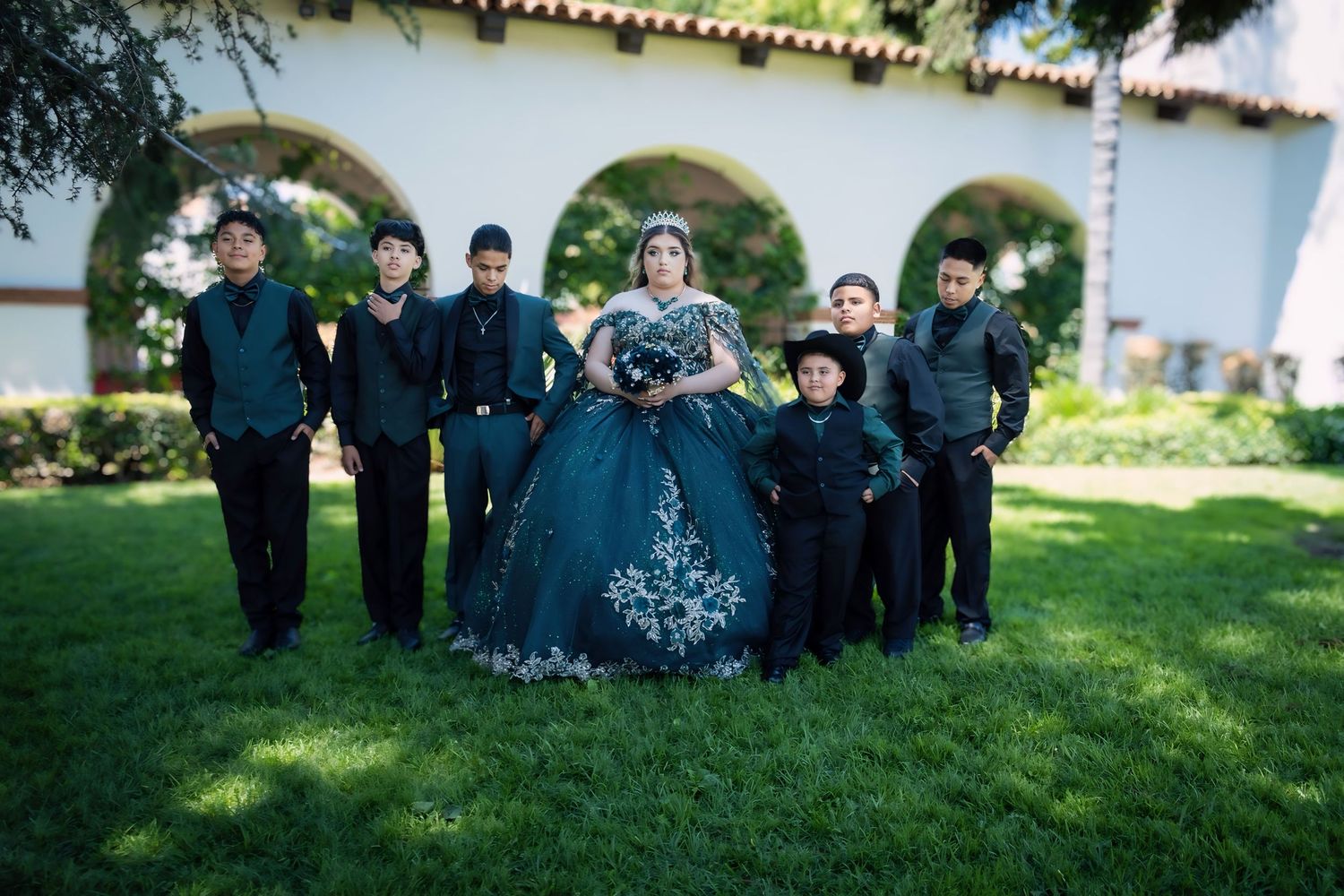 Quinceañera girl in black embroidered ball gown poses with chambelanes on green lawn.