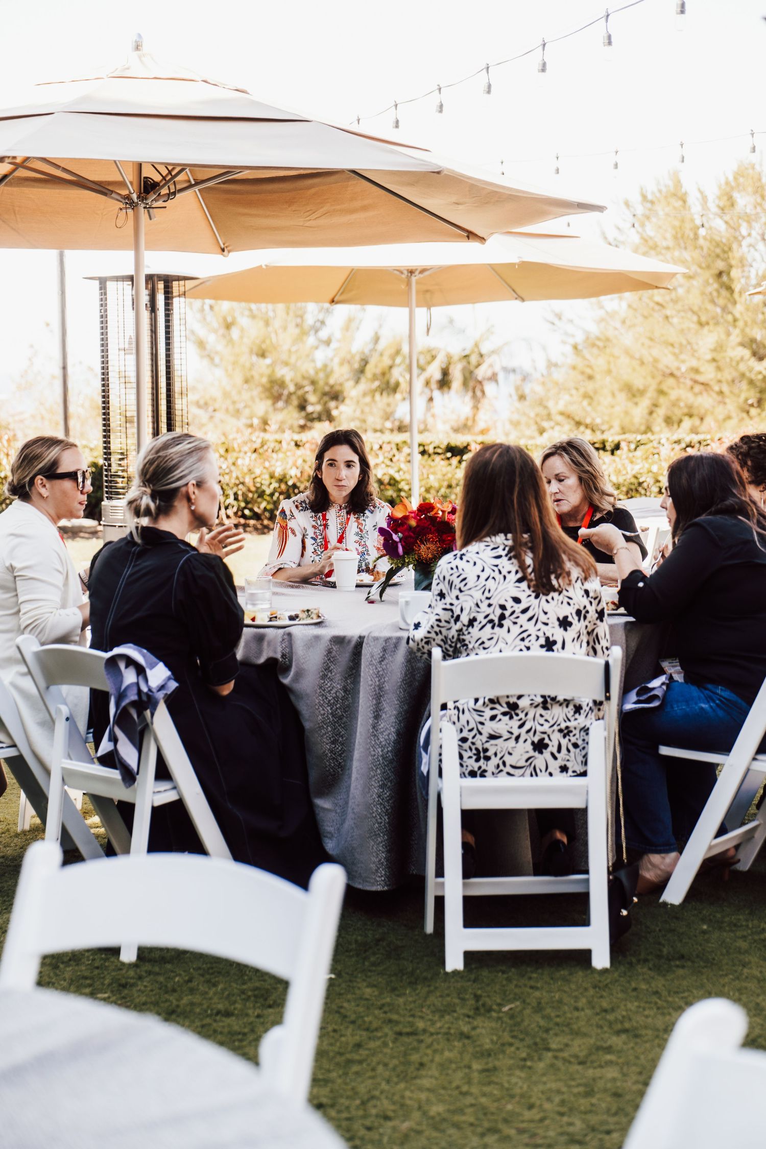 Women seated at round outdoor tables with umbrellas enjoying a garden luncheon event on green lawn.