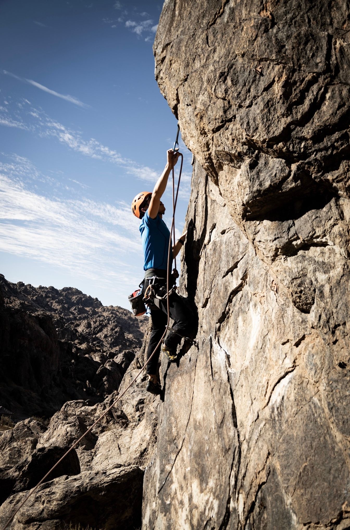 A rock climber in a blue shirt and helmet scales a steep cliff face with ropes against a blue sky.