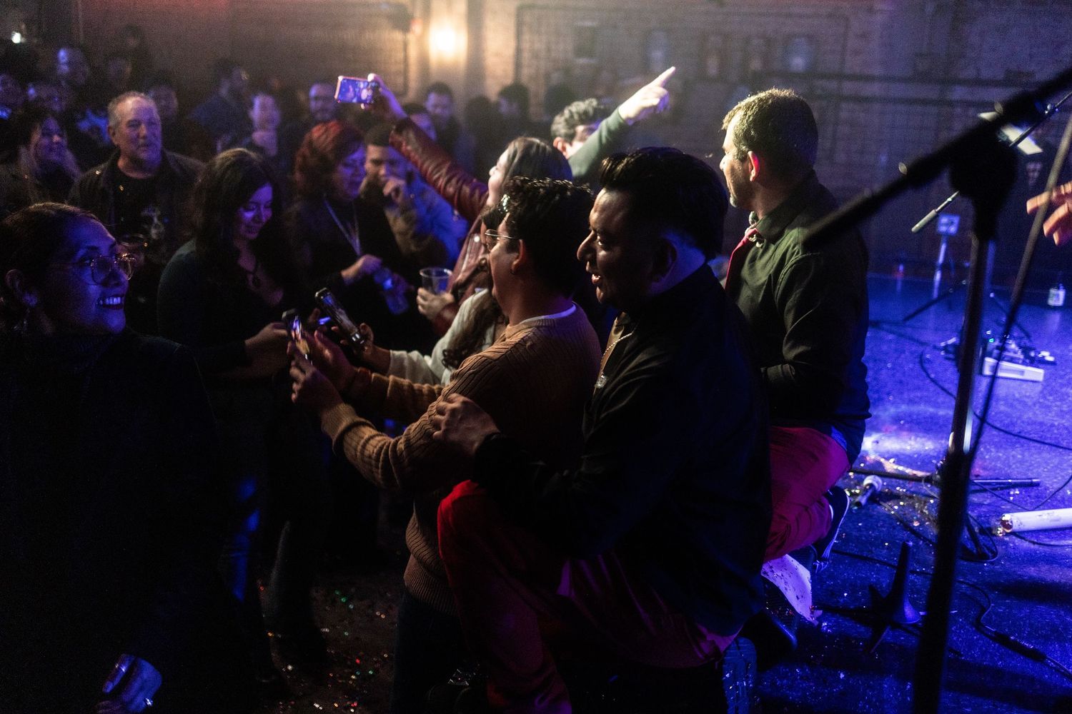 Crowd of concert attendees taking photos and dancing together under blue stage lights at a live music event.