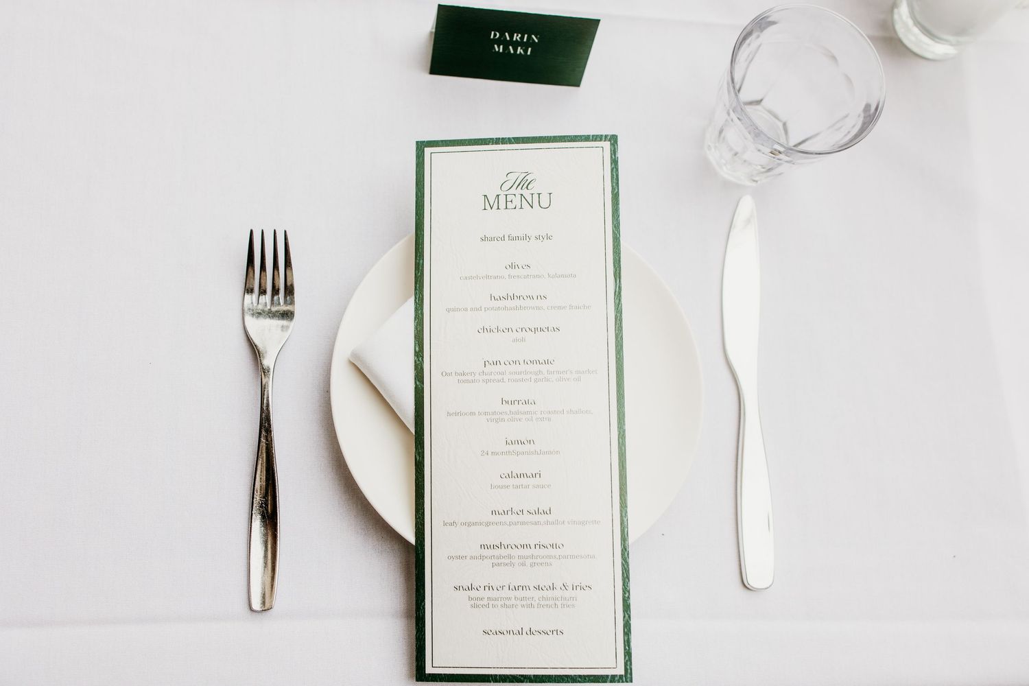 Elegant wedding place setting with a green-bordered menu card, fork, knife, and glass on a white tablecloth.