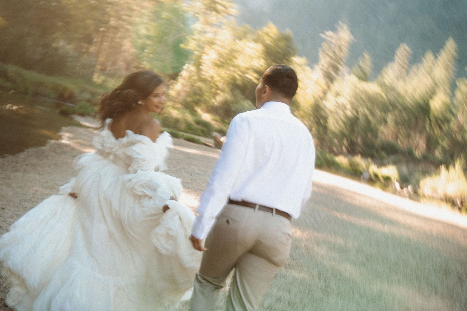 Bride and groom holding hands running outdoors in sunlit mountain setting, bride in flowing white gown.