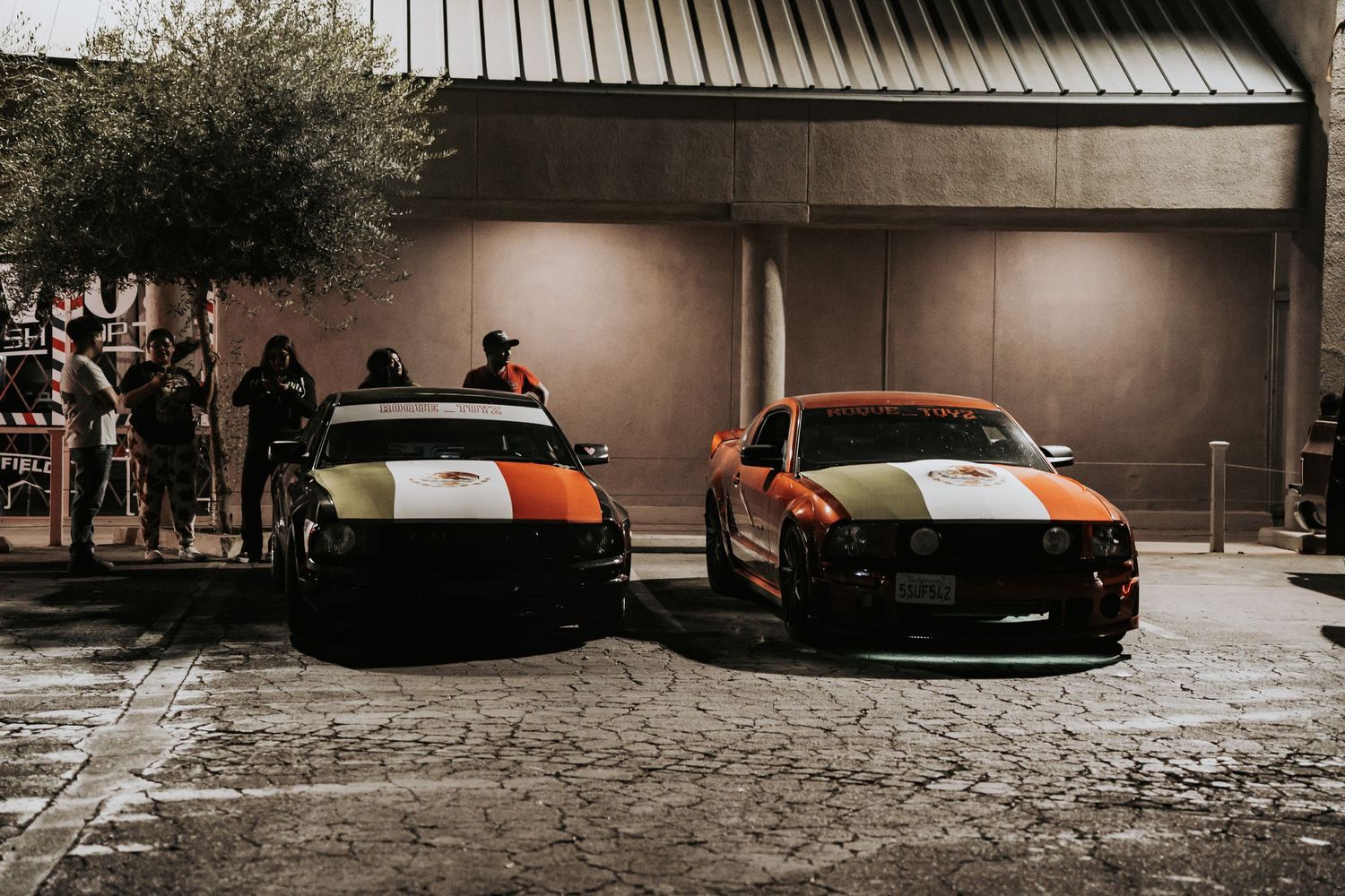 Two colorful racing-striped sports cars parked outside a building at night with spectators nearby.