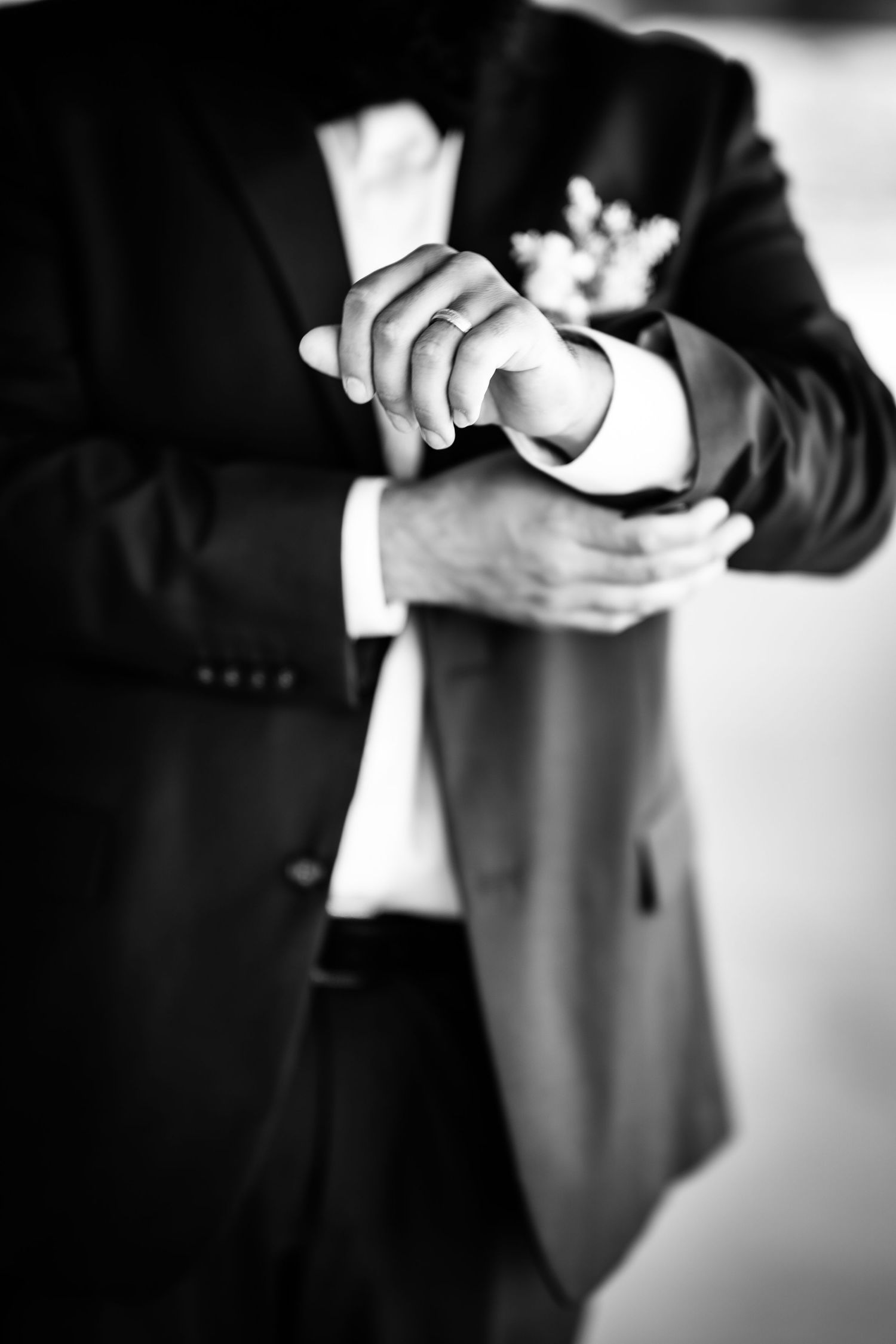 Groom adjusting his cufflinks in a black tuxedo with boutonniere, black and white wedding photo.
