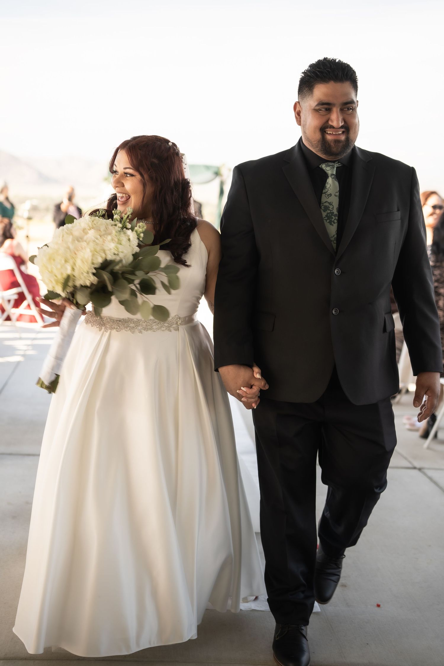 Smiling bride in white gown holding bouquet walks hand-in-hand with groom in black suit after wedding ceremony.