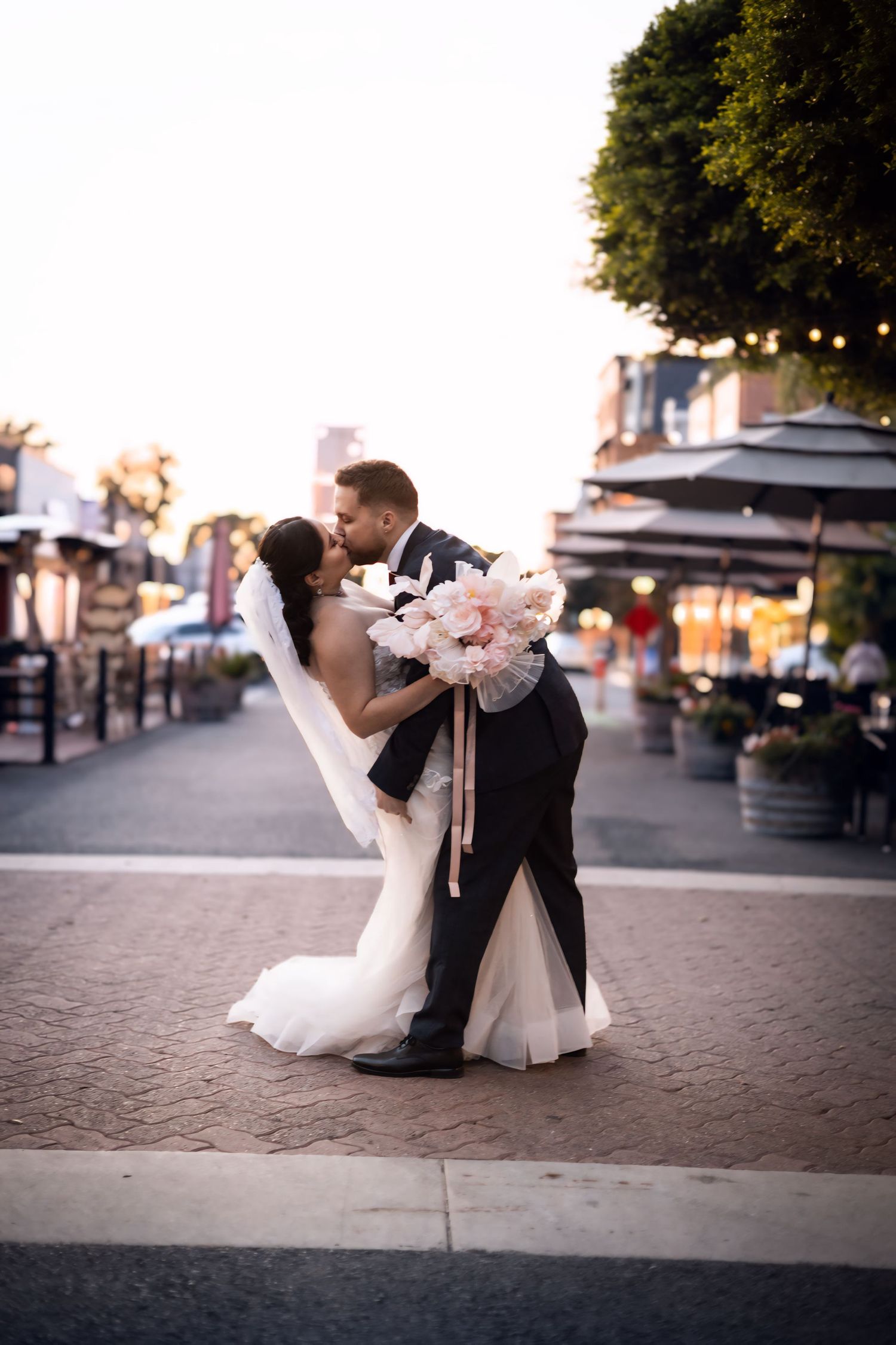 Groom lifting bride for a kiss on a city street, bride holding pink peony bouquet at sunset.