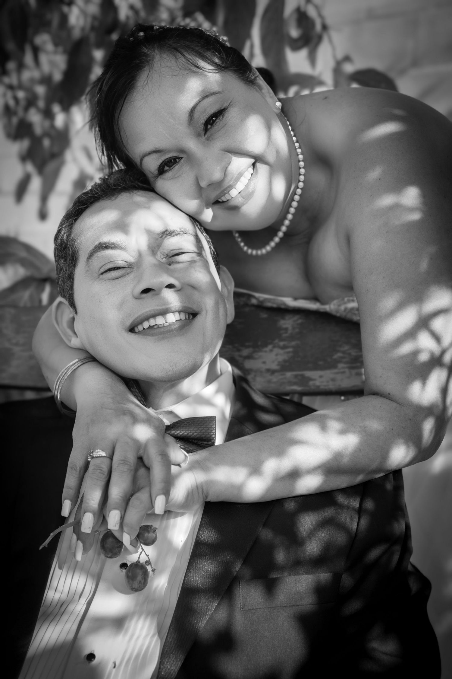 A happy couple embraces in a black and white portrait, smiling joyfully in dappled sunlight.
