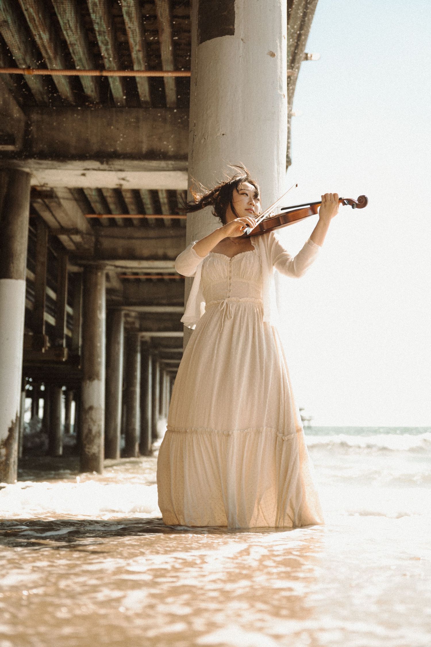 Woman in white dress playing violin under a pier with ocean waves.
