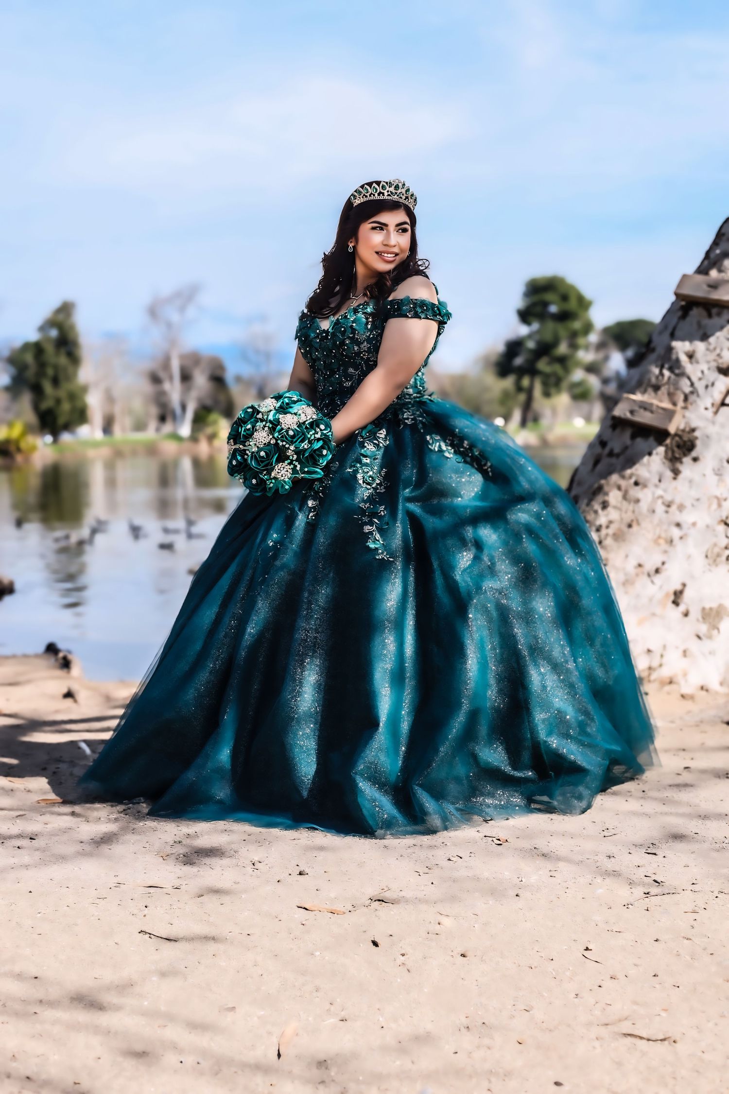 Smiling young woman in teal green quinceañera ball gown with crown holding bouquet near a lake.