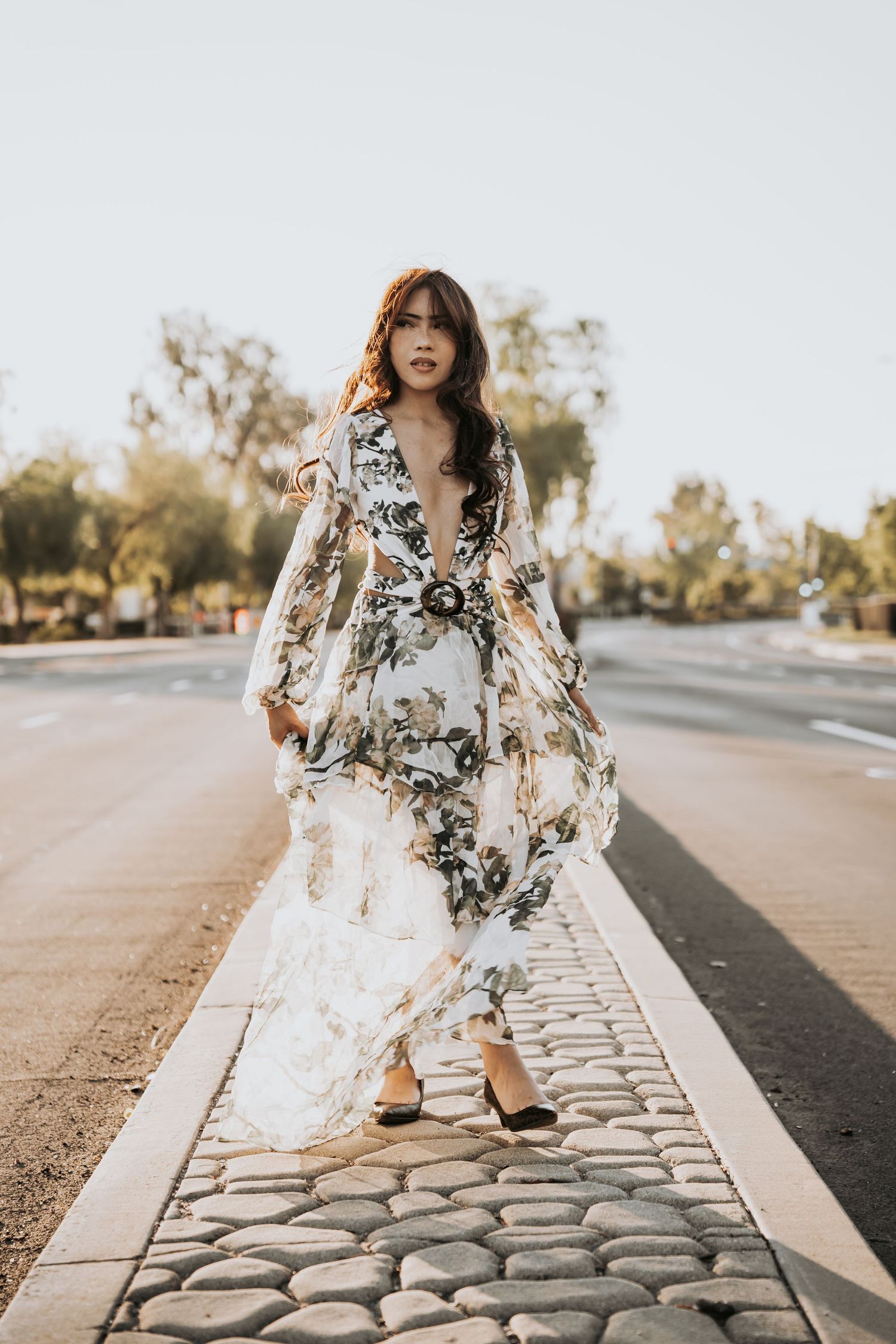 A woman in a floral long-sleeve maxi dress walking confidently along a cobblestone street curb.